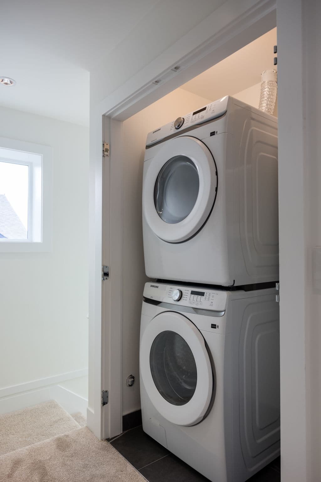 Charlton – Laundry closet with a stacked white front-load washer and dryer set behind a door, with stairs visible in the hallway.
