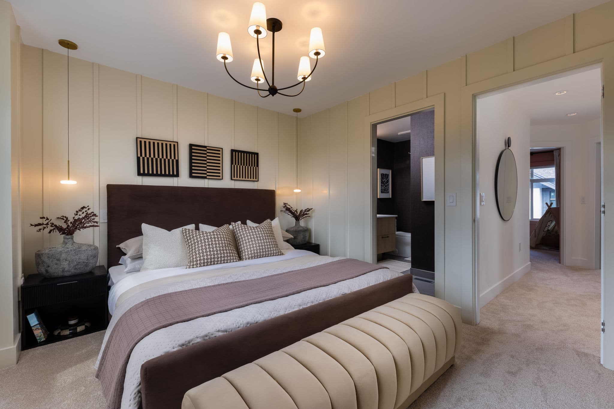 Charlton – Primary bedroom with paneled cream walls, dark headboard, chandelier, and a doorway view into an ensuite bathroom and hallway.
