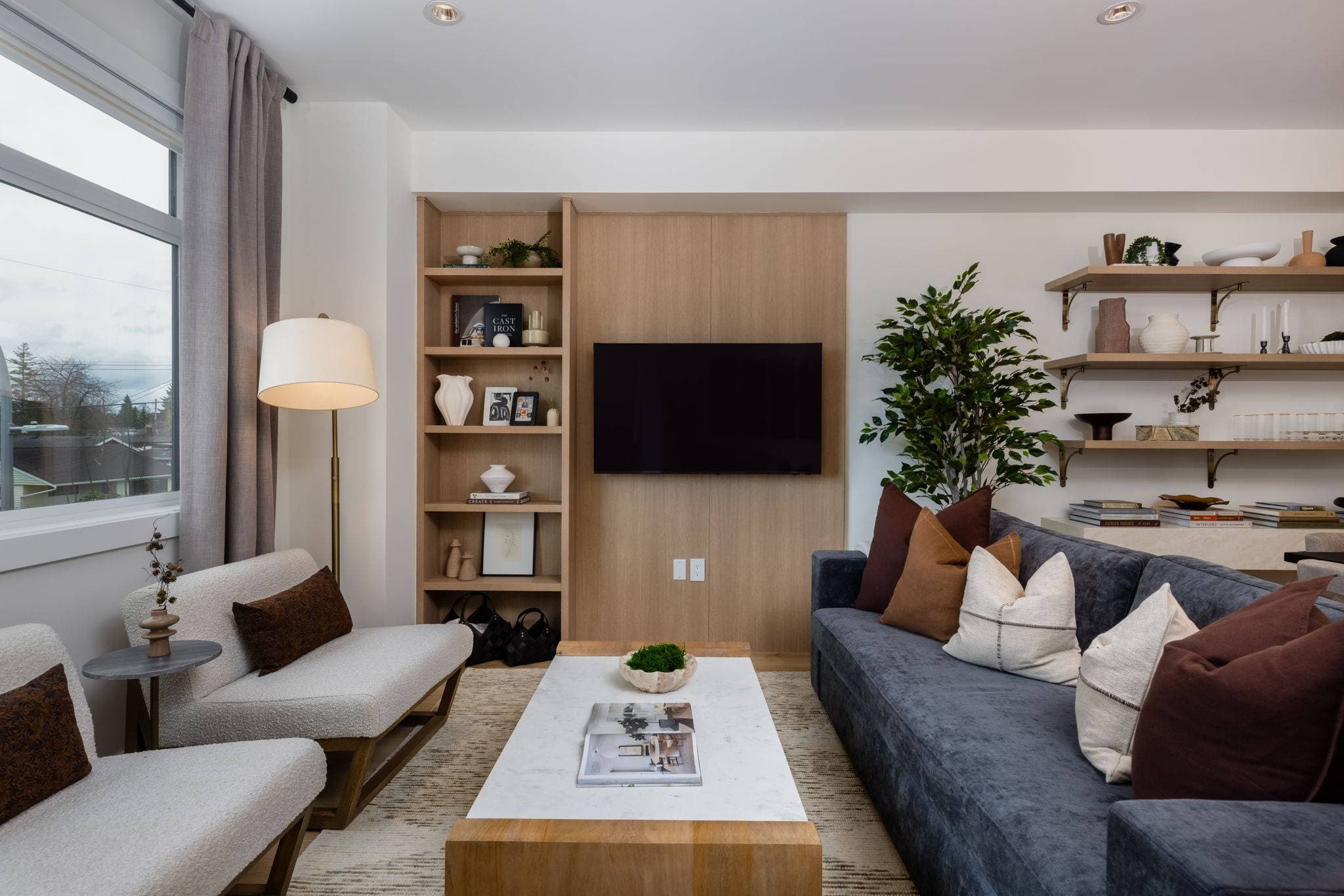 Charlton – Living room with a wood-panel TV wall, built-in shelving with decor, dark blue sofa with mixed pillows, and a floor lamp by the window.