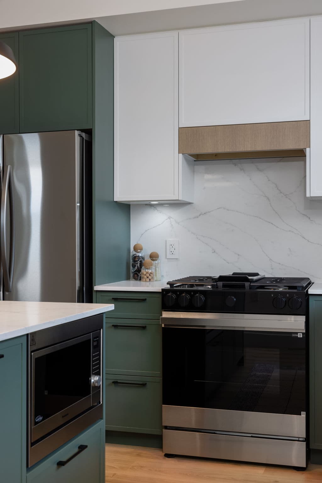 Charlton – Close-up of a kitchen gas range with marble backsplash, white and green cabinetry, and a built-in microwave in the island.