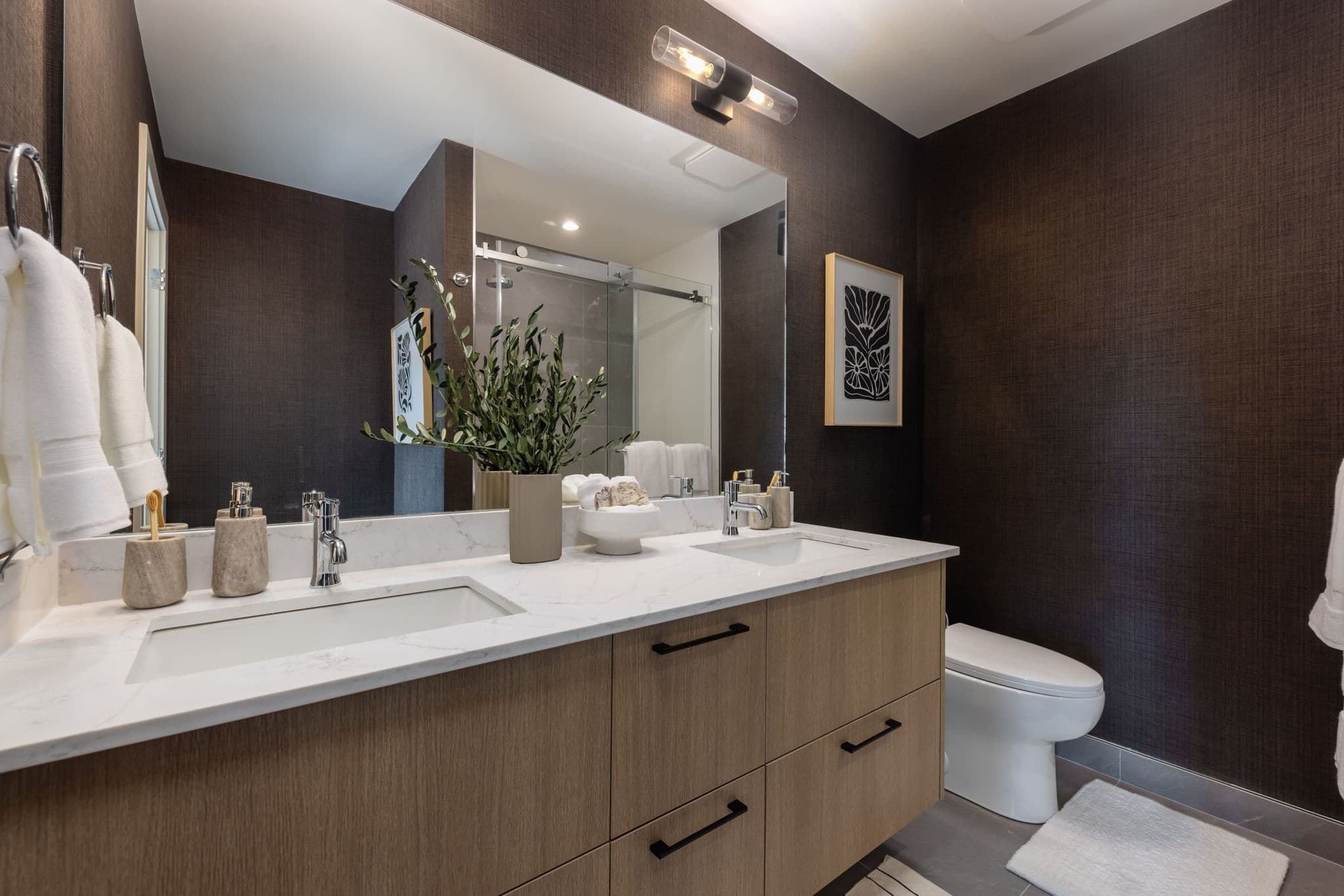Charlton – Bathroom with dark textured wallpaper, wood double vanity, glass-enclosed shower, and a black-and-white floral framed print.