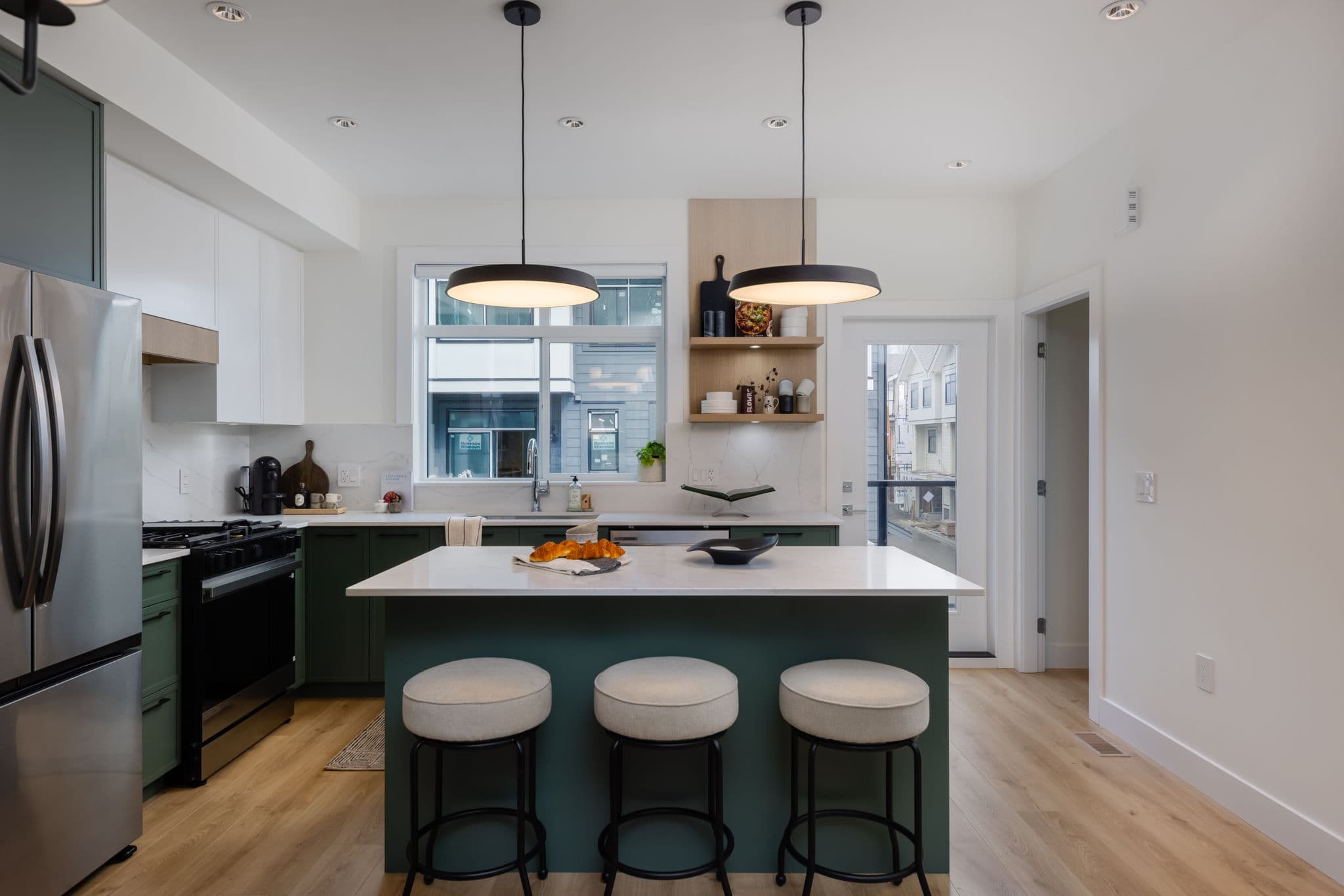 Charlton – Kitchen island with three stools under two black pendant lights, gas range and fridge in the green-cabinet run beyond.