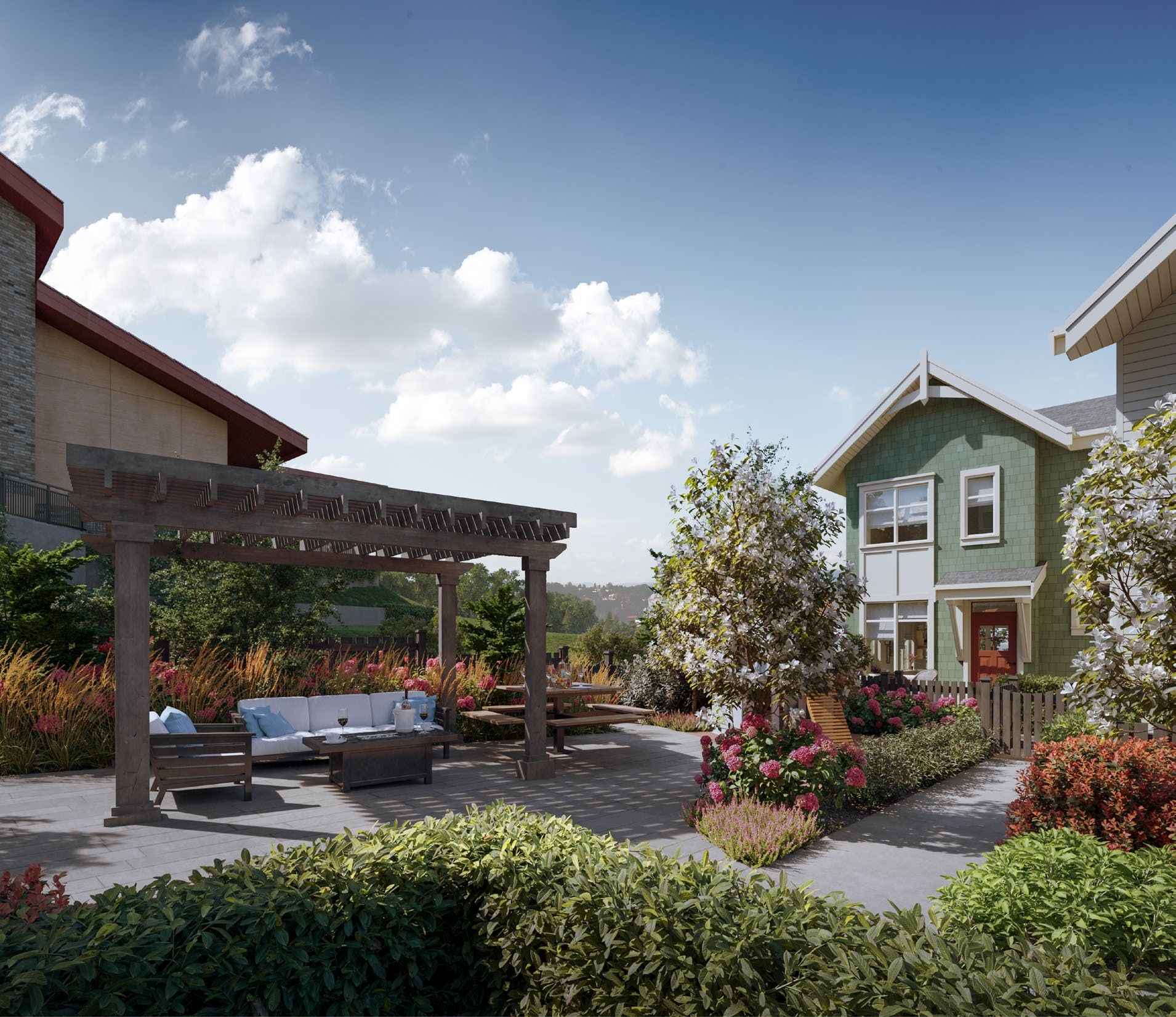 Westview Townhomes – A sunny outdoor courtyard and patio area featuring a wooden pergola over cushioned lounge seating, a coffee table, and a picnic bench. Lush landscaping with flowering trees, pink blossoms, and manicured shrubs borders the paved gathering space adjacent to green craftsman-style townhomes.