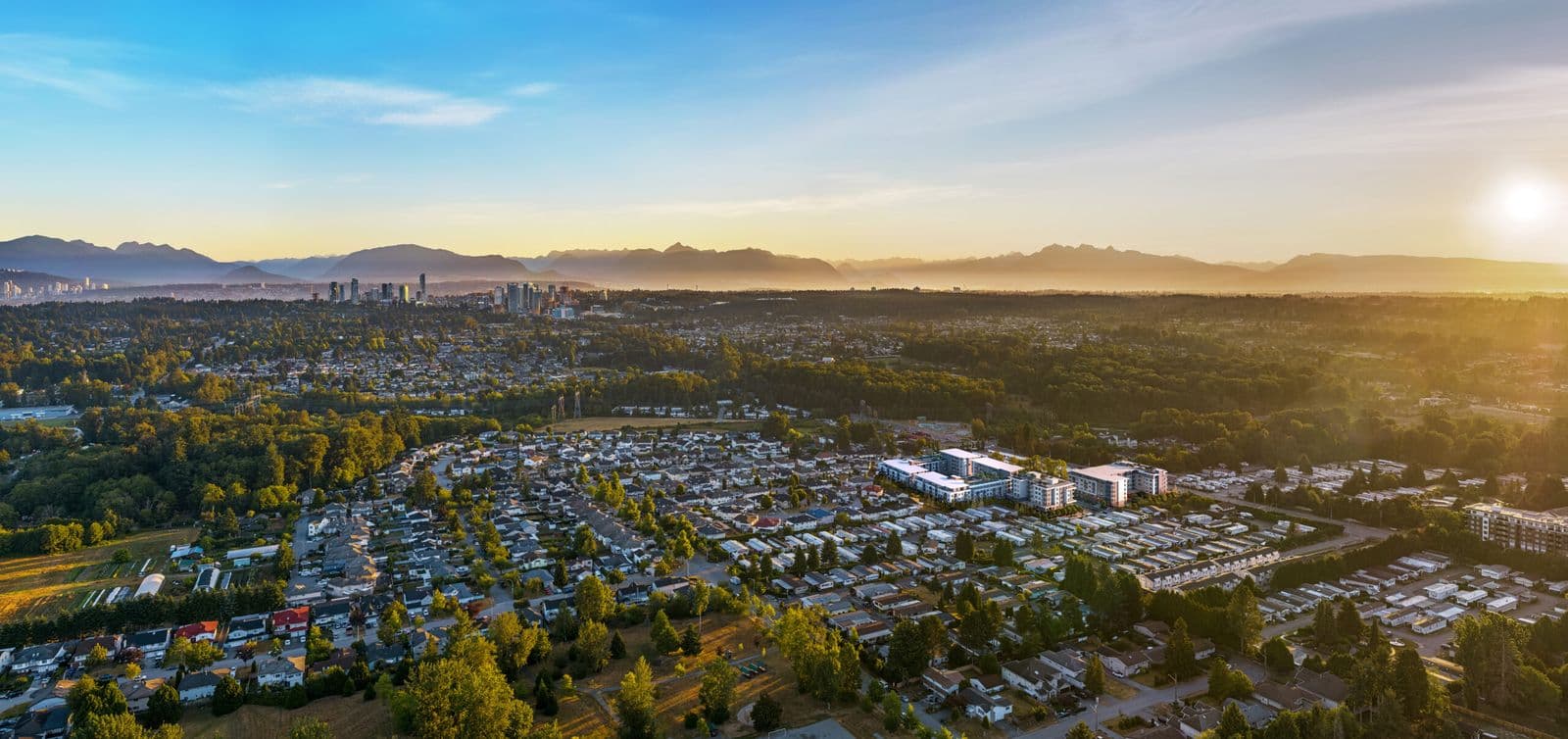 Spera – Drone view at sunrise over the surrounding low-rise suburb with the development's mid-rise buildings visible among trees, city skyline and mountains in the distance.