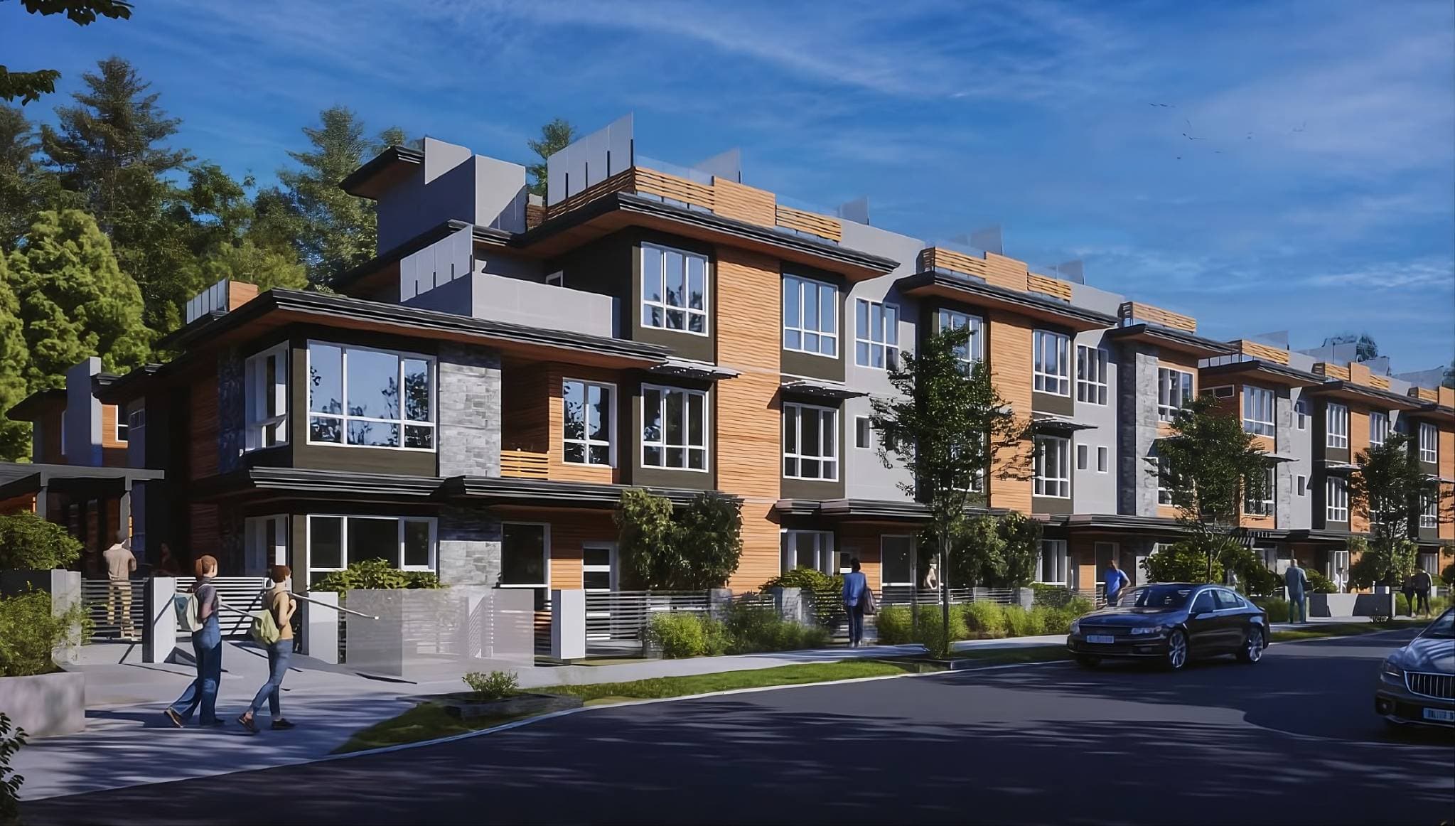 3428-3464 Mount Seymour Parkway Townhomes – A modern three-story townhouse development with warm wood siding, grey stone accents, and glass-railed rooftop patios under a clear blue sky. Pedestrians walk along the manicured sidewalk and cars are parked on the paved street in front of the lush green tree backdrop.