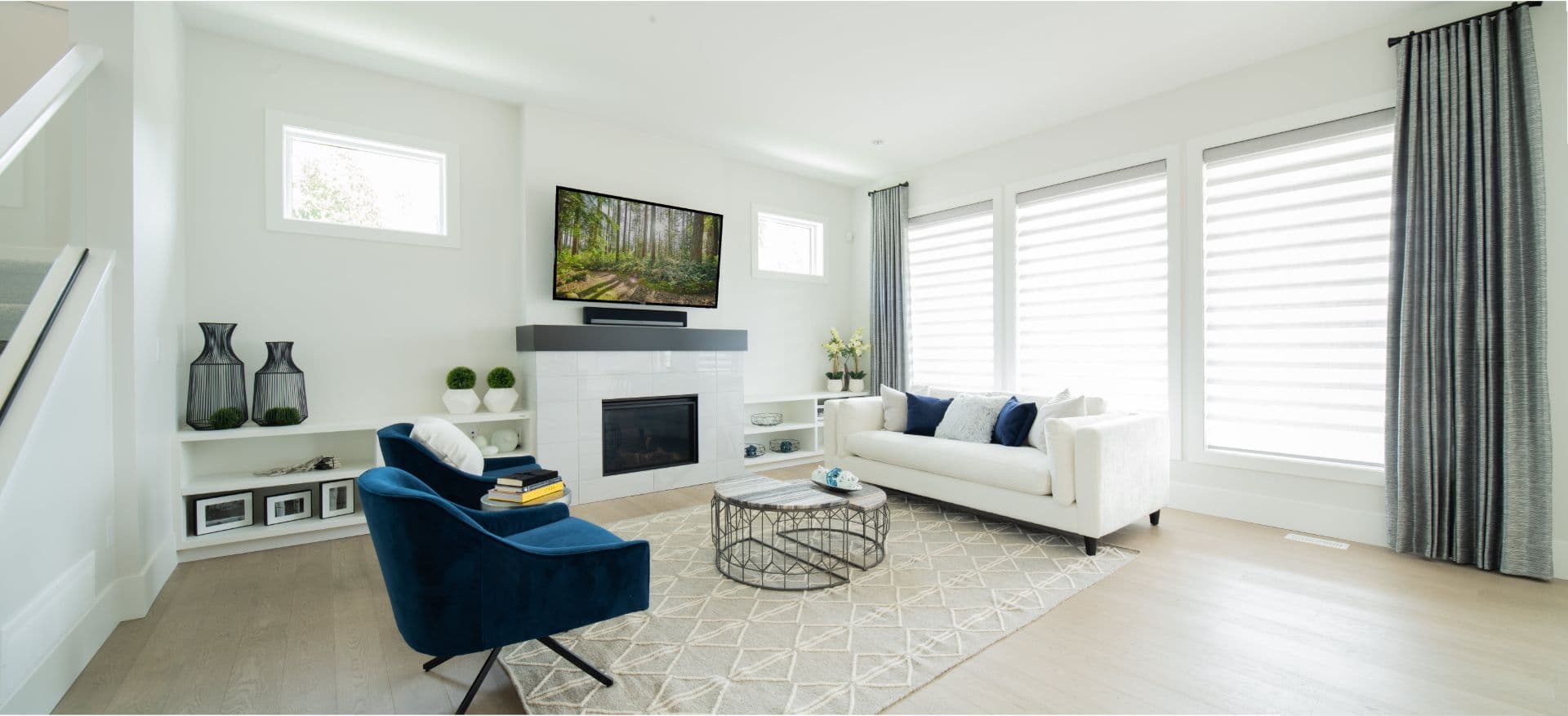 James Hill – Bright living room with a tiled fireplace, mounted TV, navy blue chairs, a white sofa, and large windows with sheer blinds.