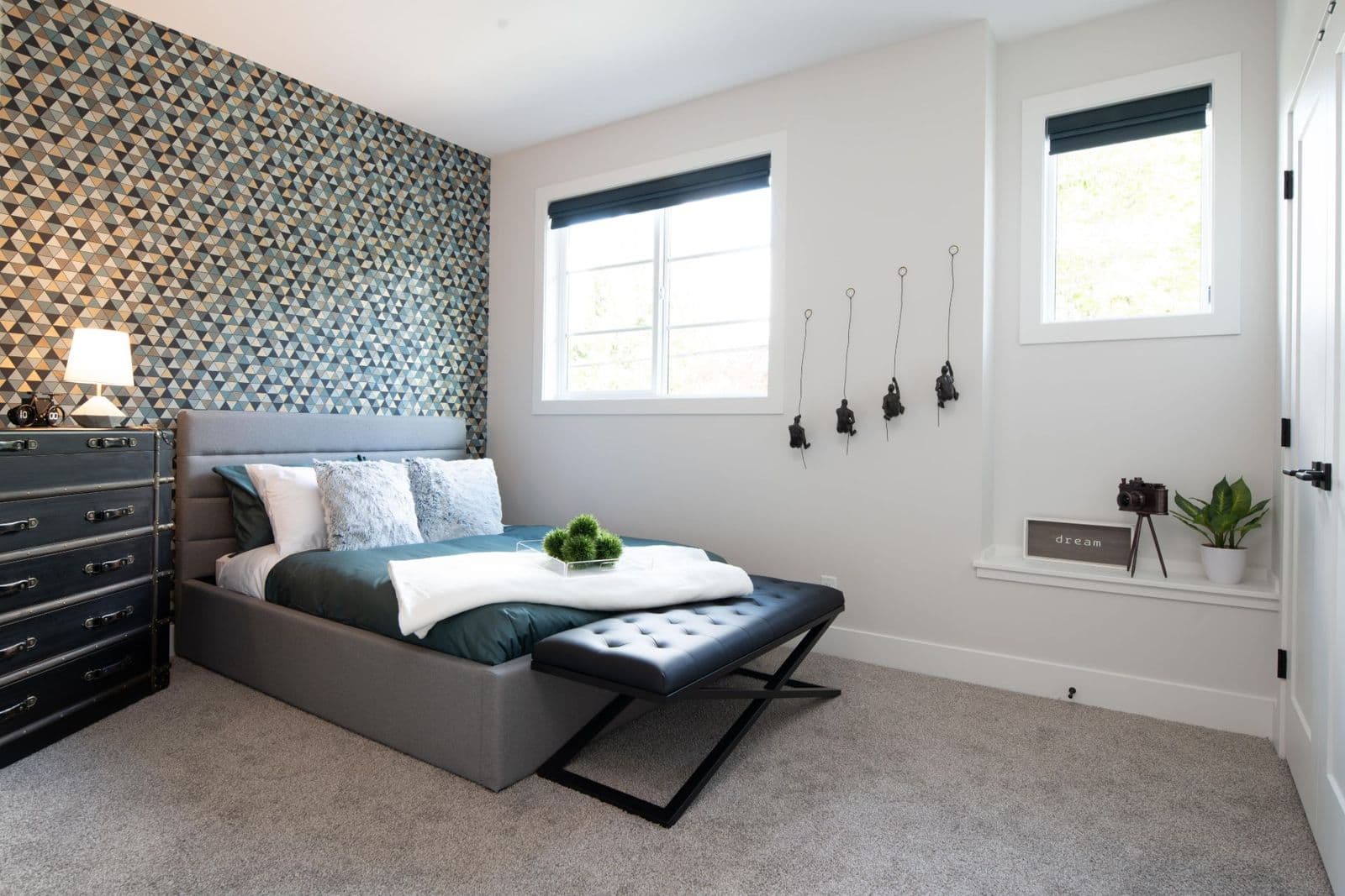 James Hill – Bedroom with a patterned blue and gold accent wall, grey upholstered bed with teal bedding, and a black tufted bench at the foot of the bed.