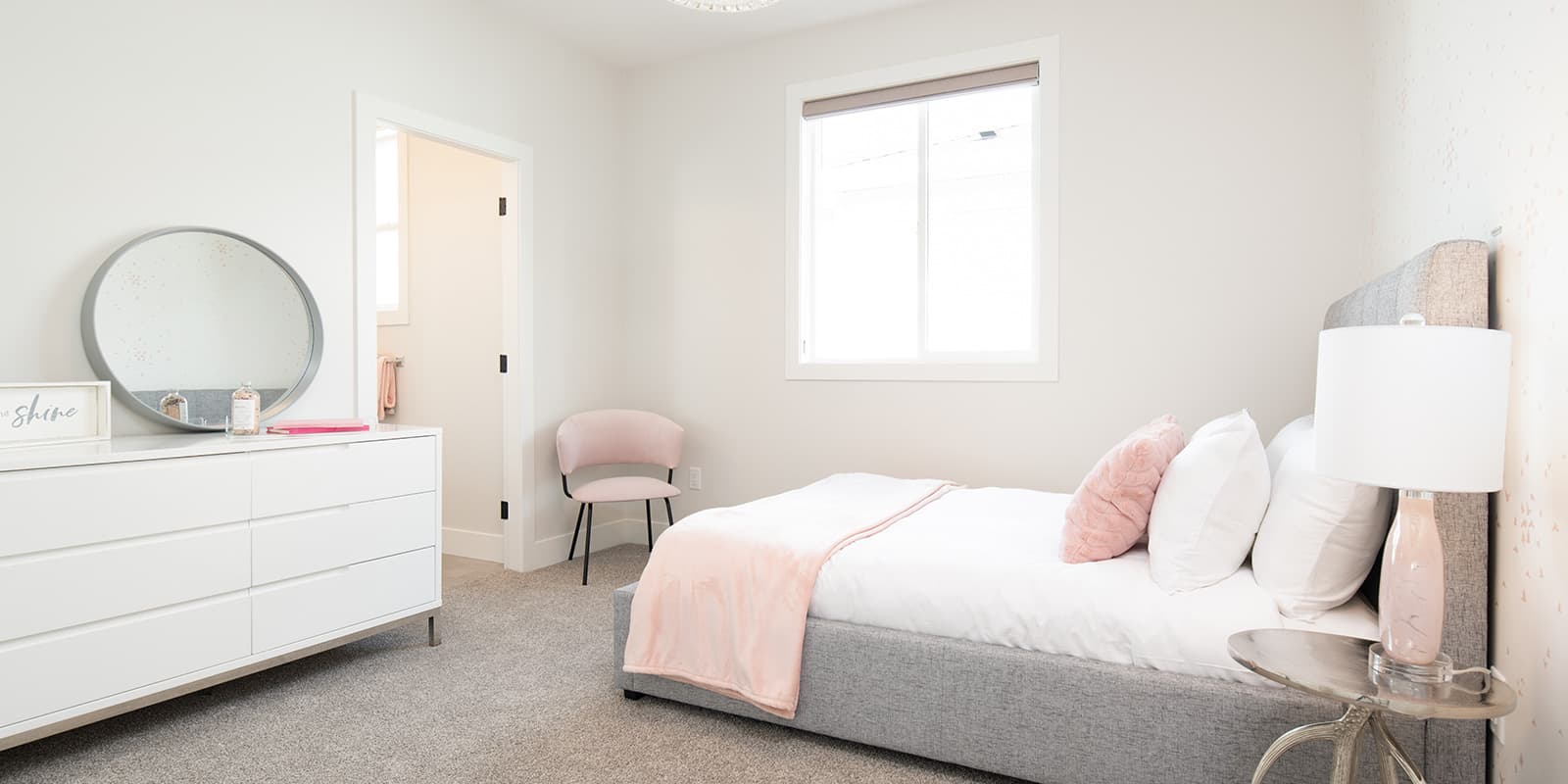 James Hill – Bedroom decorated in pink and white tones with a dresser, round mirror, small accent chair, and a bed with a pink throw blanket.