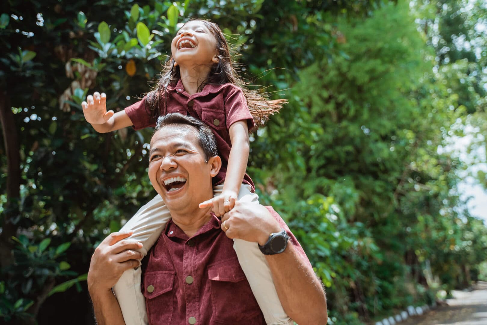 James Hill – Lifestyle stock photo of a man laughing while carrying a young girl on his shoulders outdoors among trees.