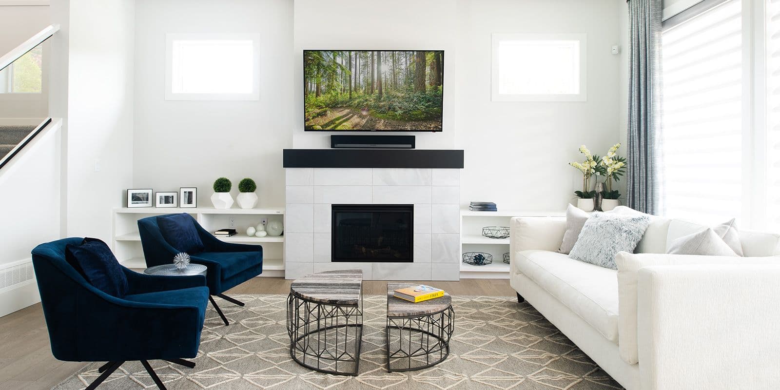 James Hill – Living room with a tiled fireplace, wall-mounted TV, two navy blue swivel chairs, a white sofa, and a patterned area rug.