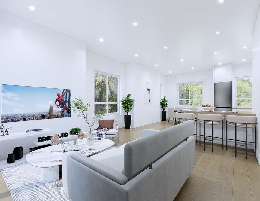 Clove – A bright, open-concept living and dining area features light wood flooring, white walls, and recessed ceiling lighting. A modern grey sofa faces a wall-mounted TV above a marble coffee table, with a kitchen island and bar stools set against windows overlooking outdoor greenery in the background.