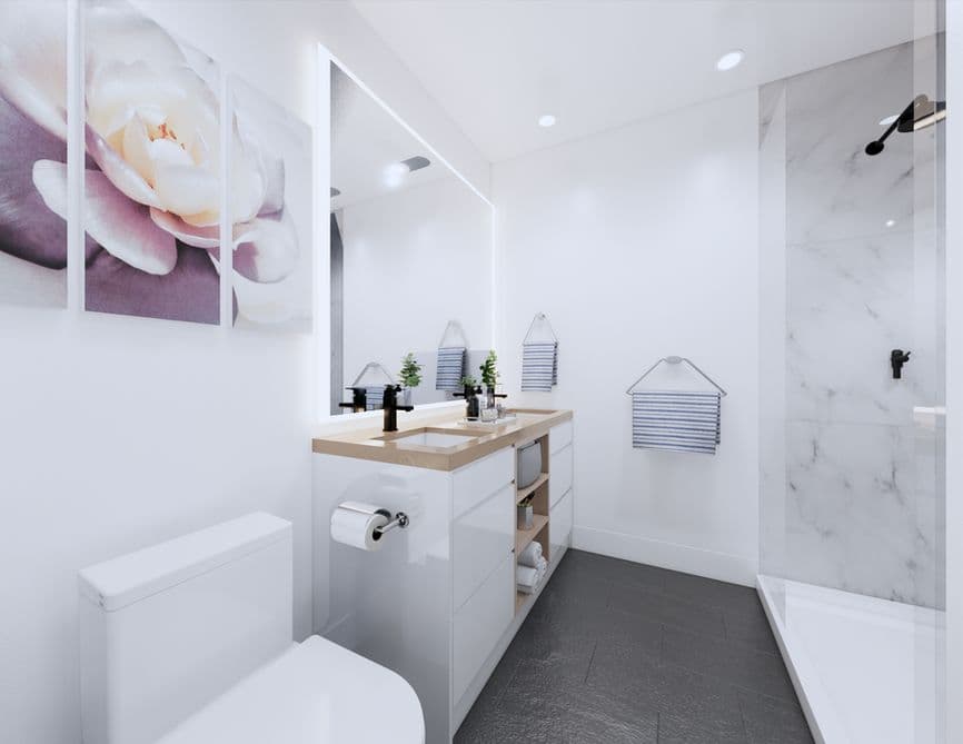 Clove – A modern, bright white bathroom featuring a sleek vanity with a wood countertop, black fixtures, and a large backlit mirror. Dark tiled flooring contrasts with white walls, a walk-in glass shower lined with marble-look tile, and a three-panel floral artwork hung beside the toilet.