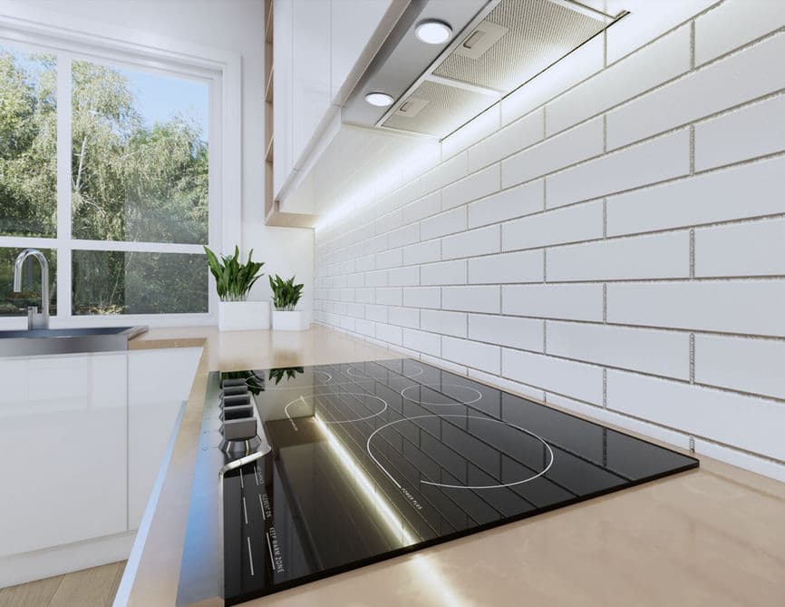 Clove – A close-up view of a modern kitchen featuring a sleek black glass stovetop set into a light countertop beneath white subway tile backsplash and an under-cabinet range hood. Natural daylight streams in from a nearby window framing green outdoor foliage.