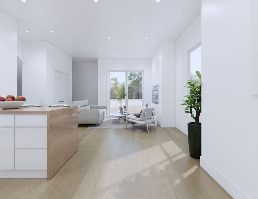 Clove – A bright, open-concept living and dining area featuring light wood flooring, white walls, and recessed ceiling lighting. A kitchen island with light stone countertops sits in the foreground, leading into a living space furnished with a modern grey sofa, an accent chair, and glass patio doors looking out toward green trees.