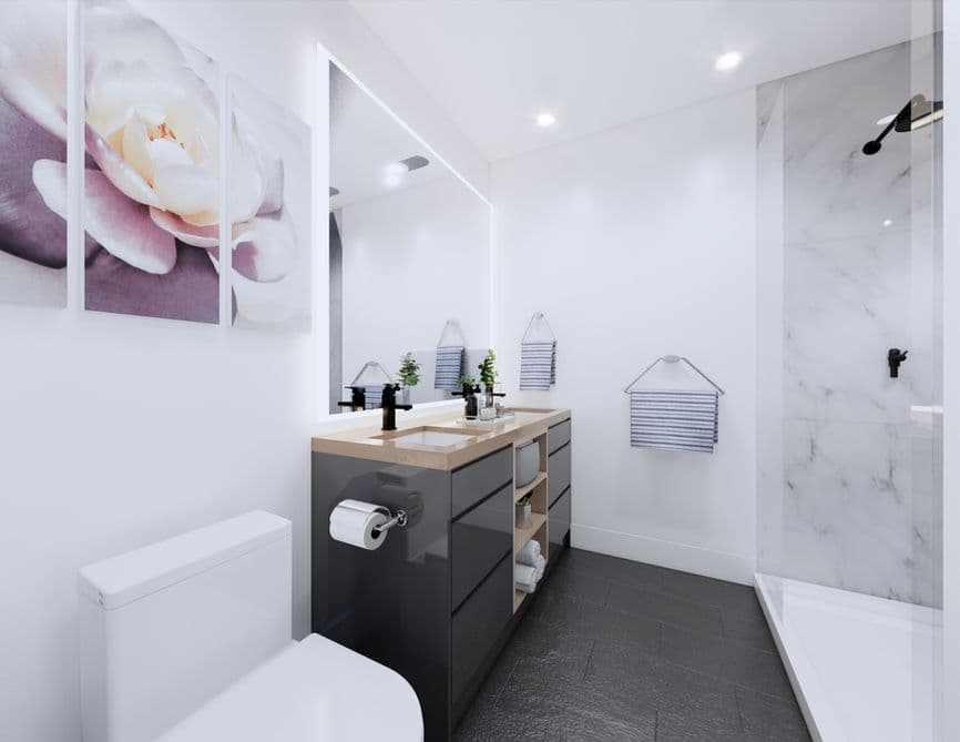 Clove – A modern, brightly lit bathroom featuring a dark grey vanity with a wooden countertop and an illuminated backlit mirror. On the right, a walk-in shower is lined with white marble-style tiles and black fixtures, complementing dark floor tiles and white walls.