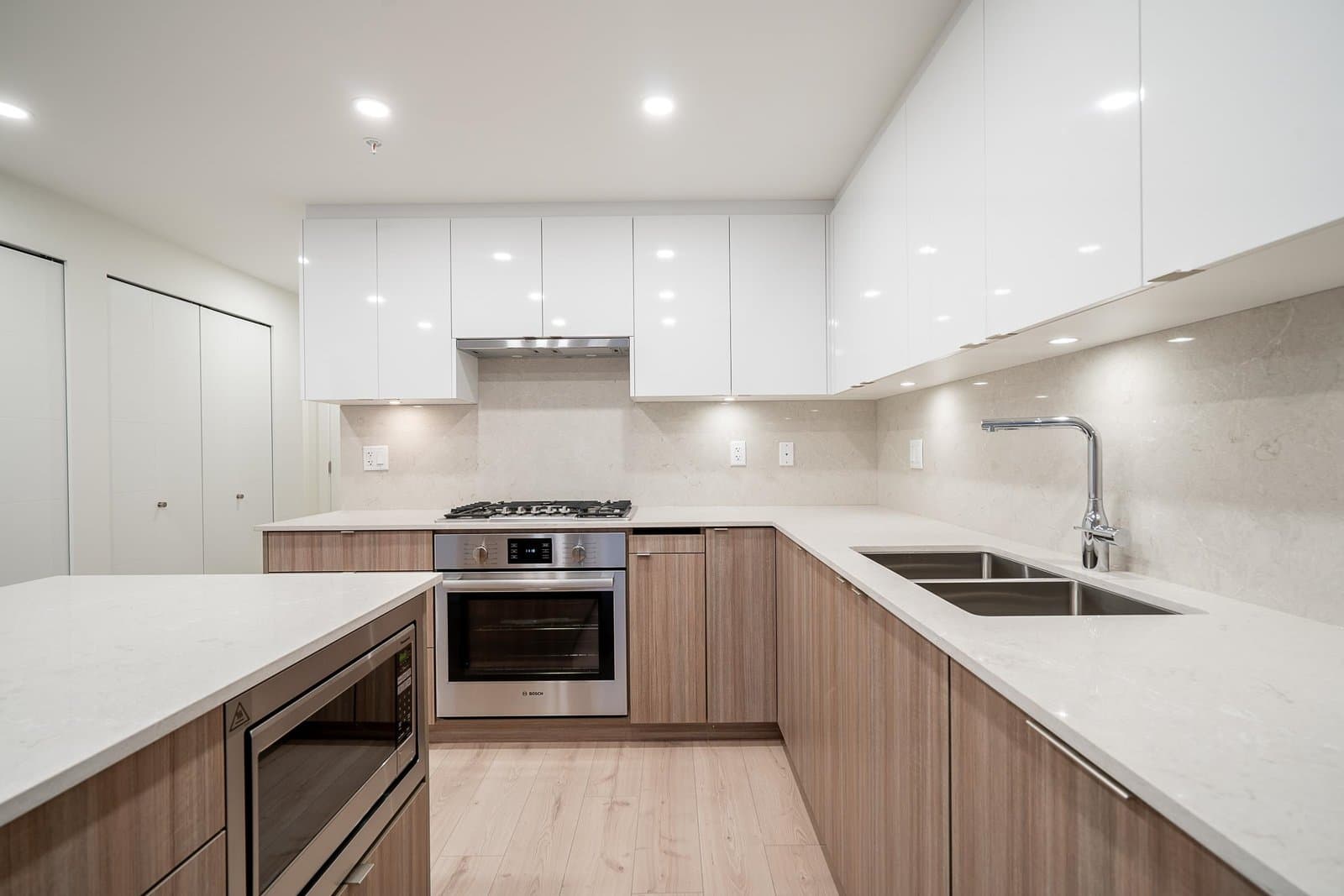 Luxio on the Park – A modern residential kitchen featuring glossy white upper cabinets, light wood grain lower cabinets, and white quartz countertops. Stainless steel appliances include an under-mount double sink with a tall gooseneck faucet, a gas cooktop with a range hood, and a built-in oven.