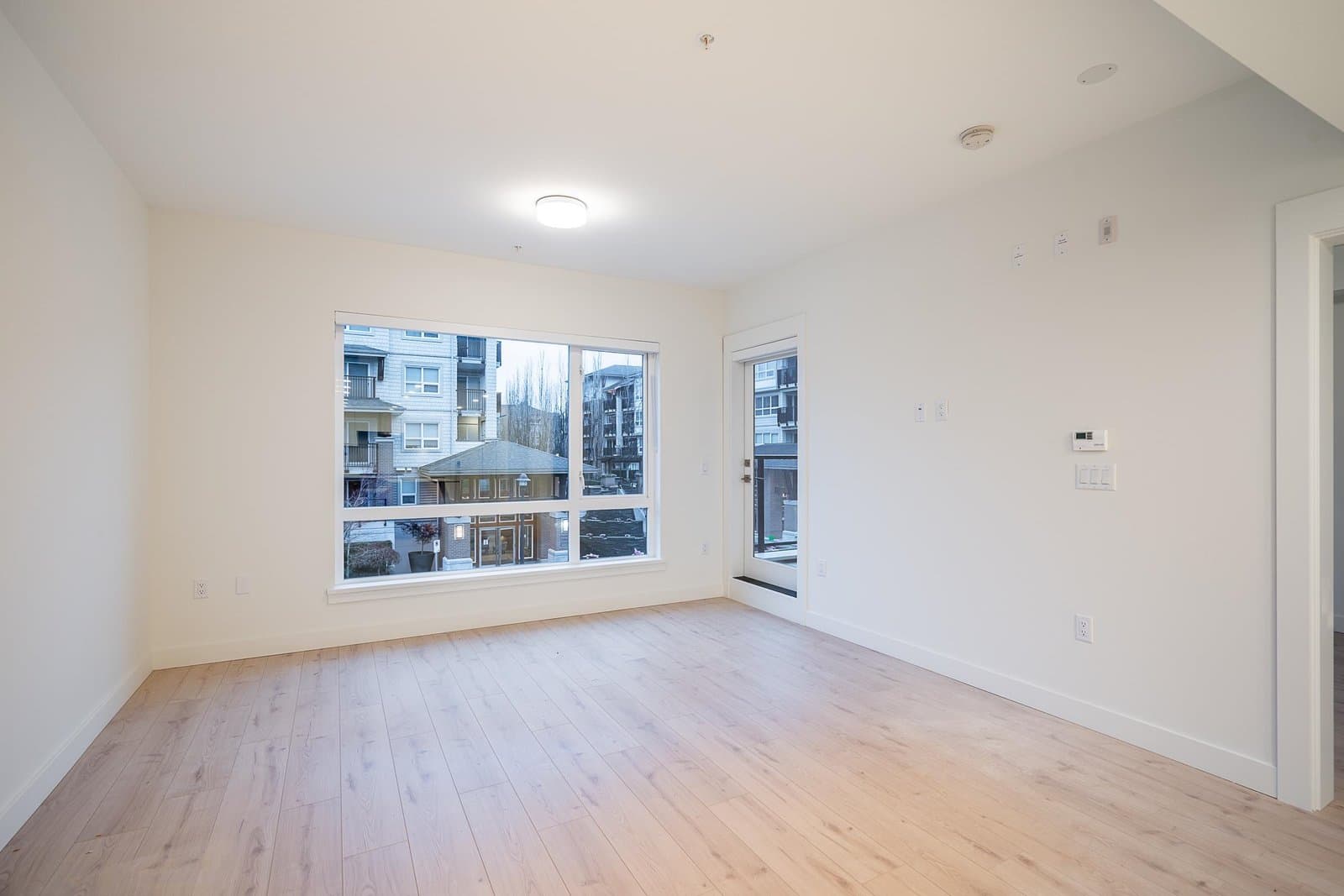 Luxio on the Park – An empty, brightly lit room features light wood laminate flooring and clean white walls. A large window and adjacent glass patio door look out onto a neighboring residential building complex during daylight.