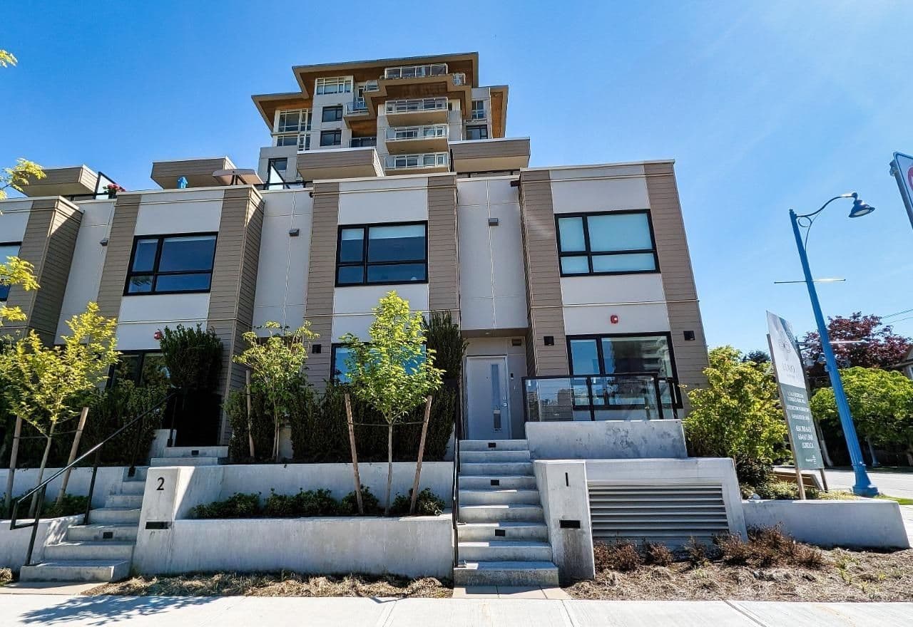 Luxio on the Park – A ground-level view looking up at a modern mid-rise residential building featuring a mix of beige siding, white stucco, and concrete staircases leading to private entrances. Small, newly planted trees and landscaped terraced planters line the front sidewalk under a clear blue sky.