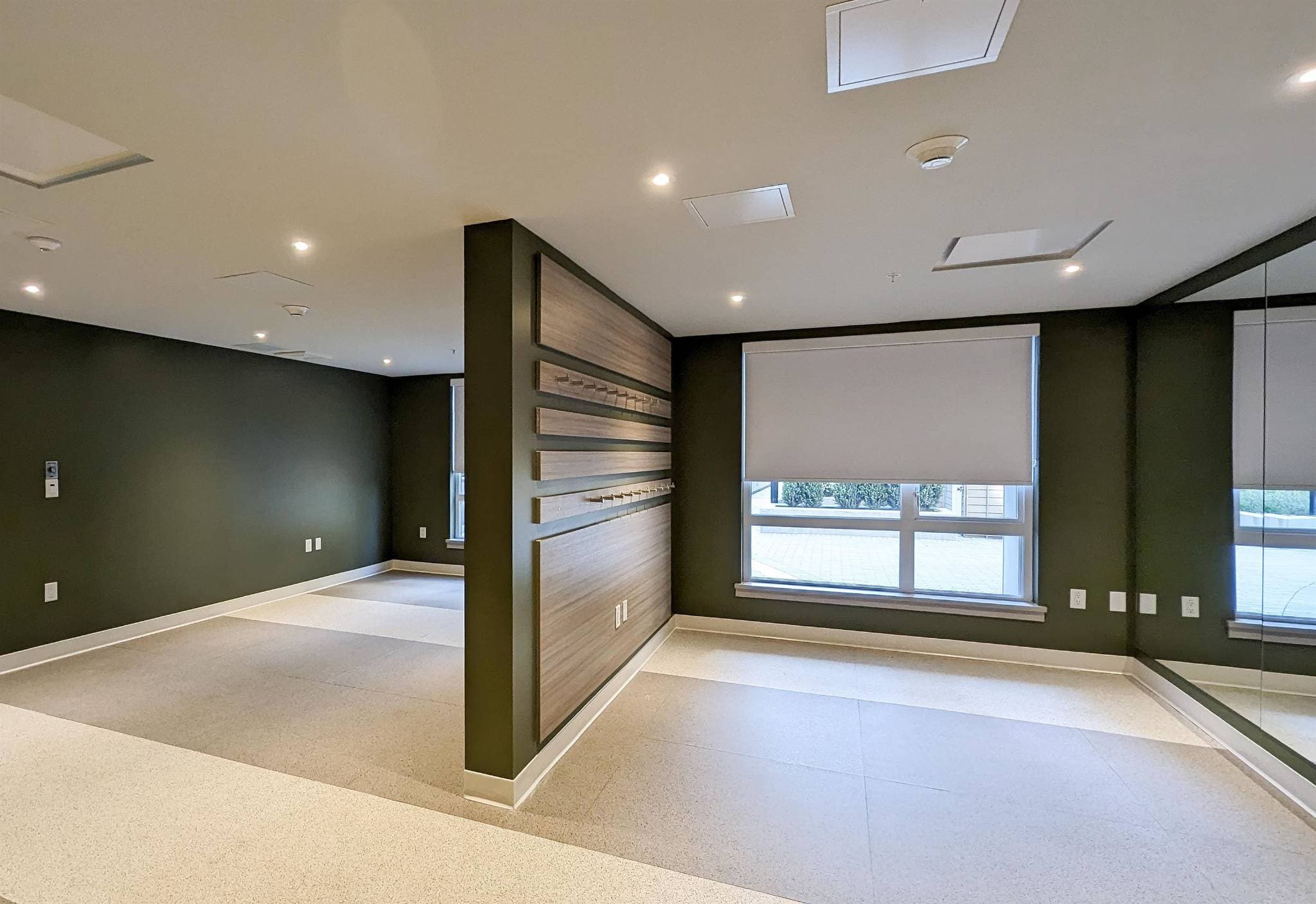 Luxio on the Park – A modern, empty indoor fitness or studio space featuring deep olive green walls, light beige tiled flooring, and a large wall mirror on the right. A central dividing wall is fitted with horizontal wood-paneled accent slates with wall hooks, next to a large window covered by a white roller shade that lets in bright daytime light.