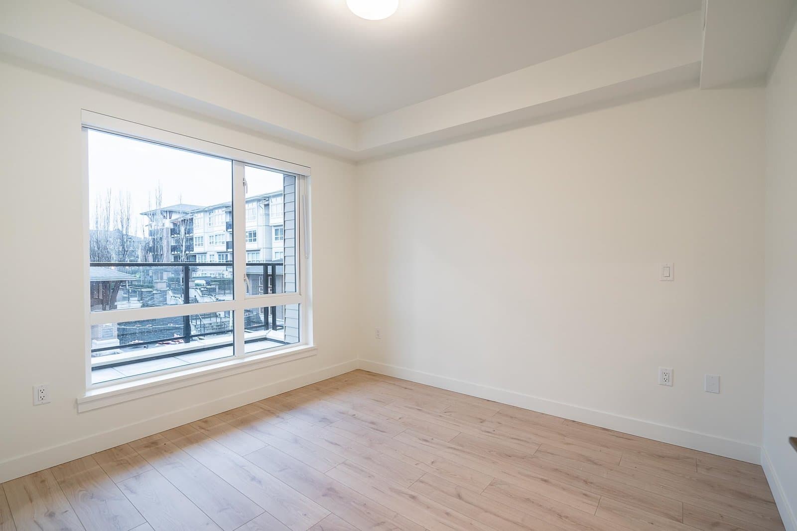 Luxio on the Park – An empty, bright room features light wood-look flooring and crisp white walls illuminated by natural light. A large window looks out onto a glass-railed balcony and neighboring residential buildings under a bright daylight sky.