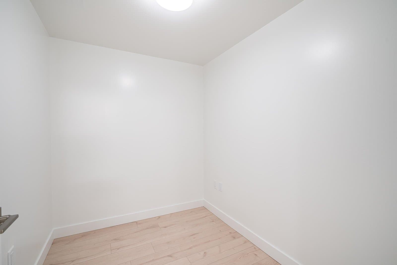 Luxio on the Park – An empty interior room featuring freshly painted white walls, clean white baseboards, and light-toned wood laminate flooring. Soft, neutral overhead lighting illuminates the vacant nook or small bedroom space.