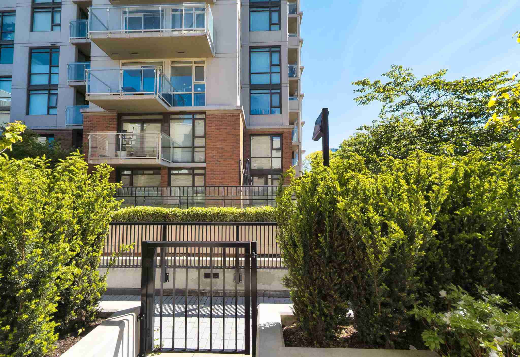 Luxio on the Park – A ground-level view of a multi-story residential building featuring glass balconies and a brick podium facade. In the foreground, lush green bushes and a black metal security gate frame the entrance area under a clear blue sky.