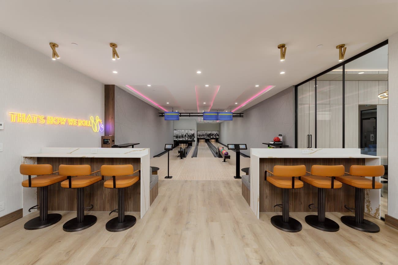 Gilmore Place Condos – Bowling alley amenity space with two lanes, orange leather stools at high tables, and a neon sign reading "That's how we roll".