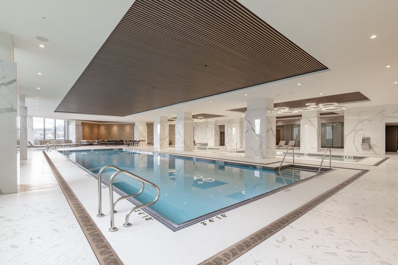 Gilmore Place Condos – Indoor swimming pool with a connected hot tub, marble columns, lounge chairs, and floor-to-ceiling windows.