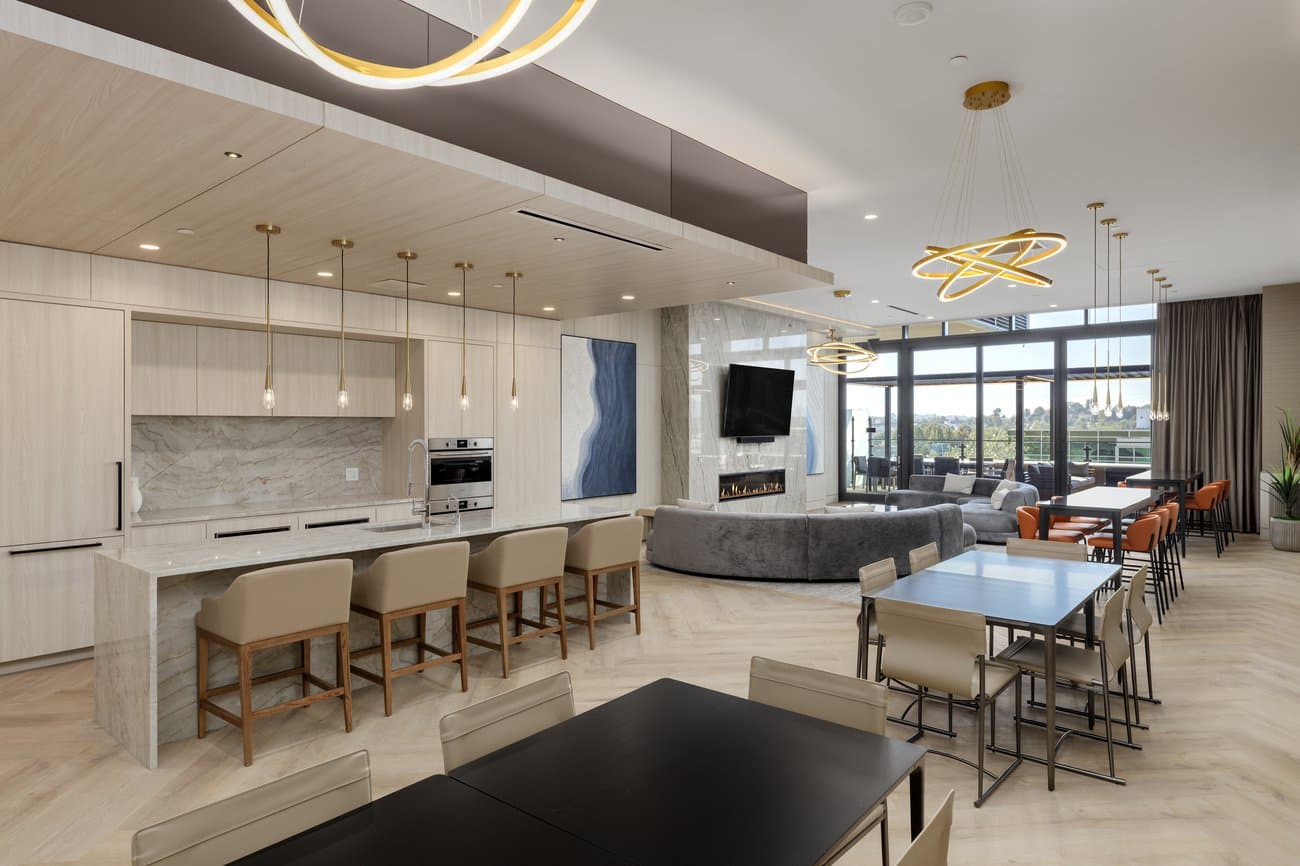 Gilmore Place Condos – Resident lounge with a kitchen island, gray sectional sofa facing a fireplace and mounted TV, and several dining tables with orange chairs.