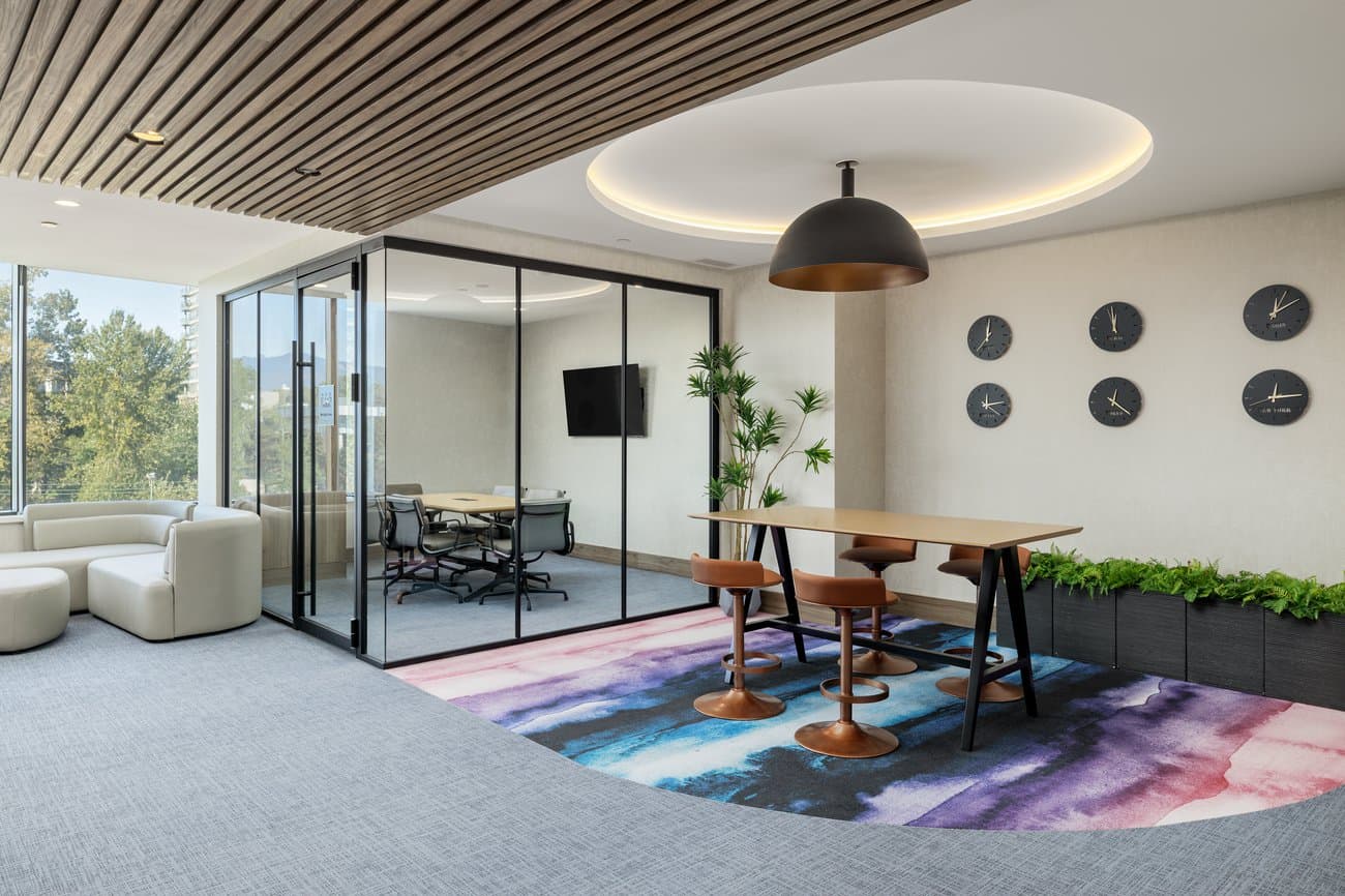 Gilmore Place Condos – Co-working lounge with a glass-enclosed meeting room, a row of decorative wall clocks, high stool seating, and a colorful area rug.
