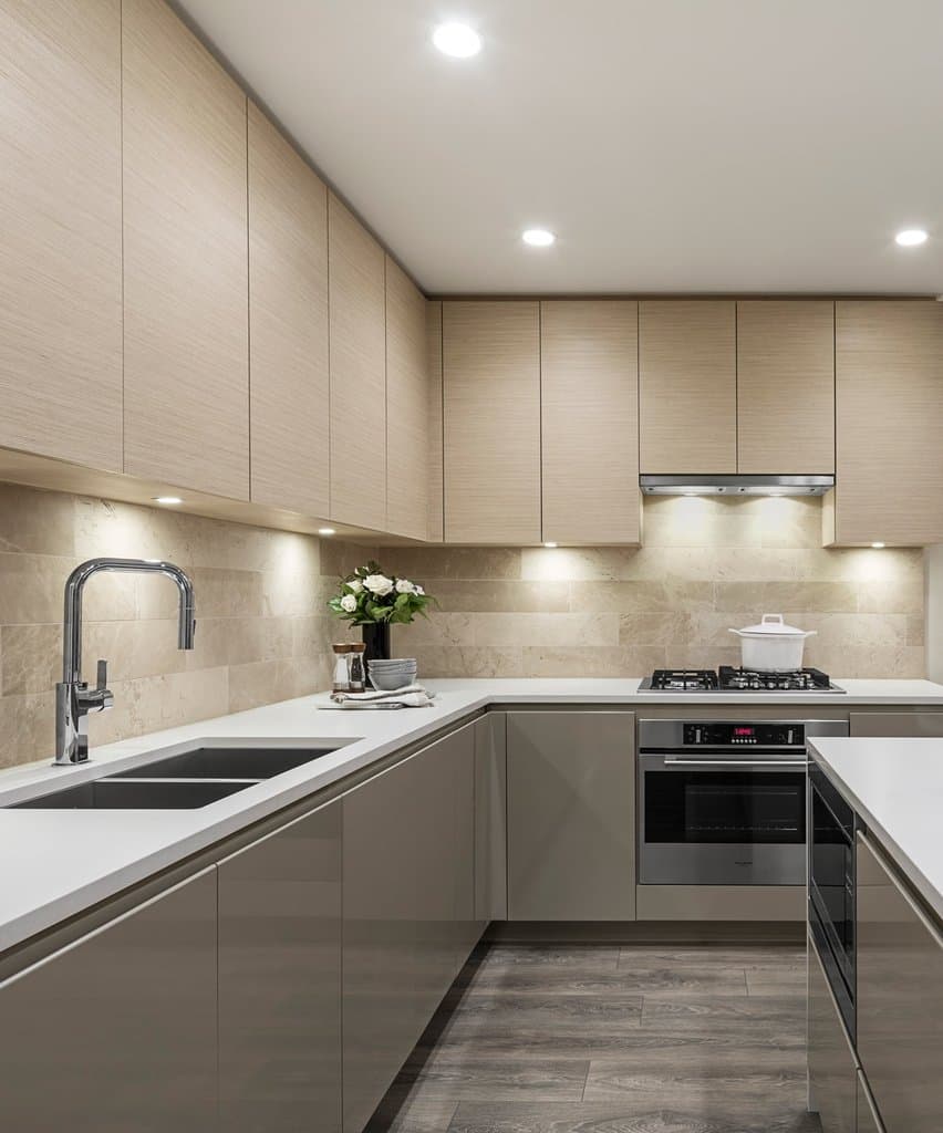 Gilmore Place Condos – Kitchen with light wood-tone cabinetry, stone tile backsplash, stainless oven, and dark wood flooring in a show suite.