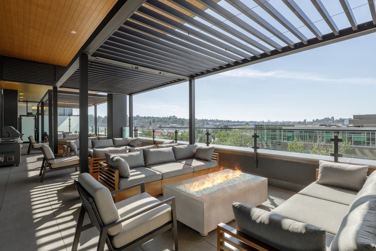 Gilmore Place Condos – Rooftop terrace lounge with gray outdoor sofas around a lit fire table, a BBQ grill, and a pergola cover overlooking the city.