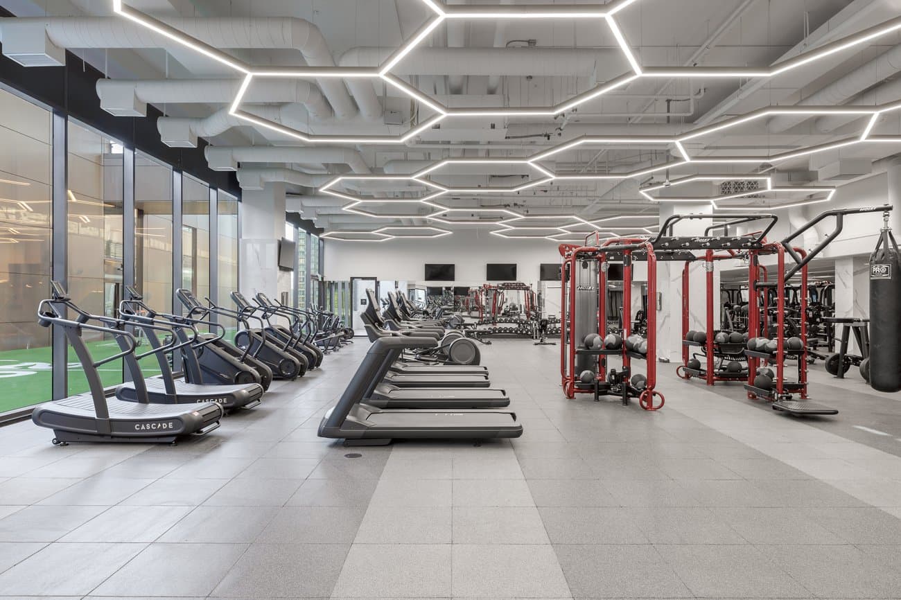 Gilmore Place Condos – Fitness gym with a row of treadmills, cable weight machines, kettlebells, and a boxing bag, lit by hexagonal ceiling light fixtures.