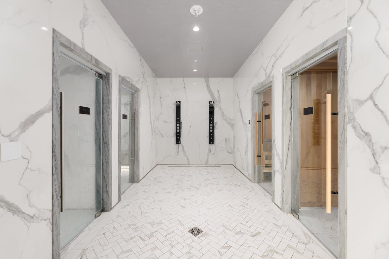 Gilmore Place Condos – Spa hallway finished in white marble with glass-enclosed steam room and sauna doors on either side and shower panels along the back wall.
