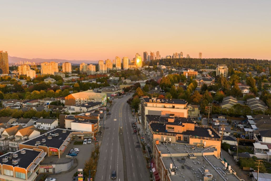 Orchard – An elevated view looking down a wide urban street flanked by low-rise commercial buildings, residential rooftops, and autumn foliage. In the distance, a skyline of high-rise towers and distant hills is warmly lit by the golden glow of a sunset.
