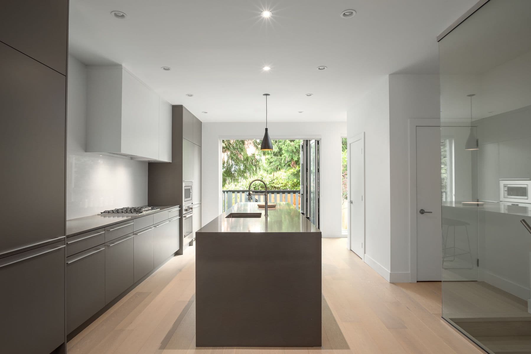 The Park Residences – A modern, minimalist kitchen features a large grey waterfall island, sleek flat-panel cabinetry in grey and white, and light hardwood flooring. In the background, open folding glass doors lead to an outdoor balcony surrounded by lush green trees.