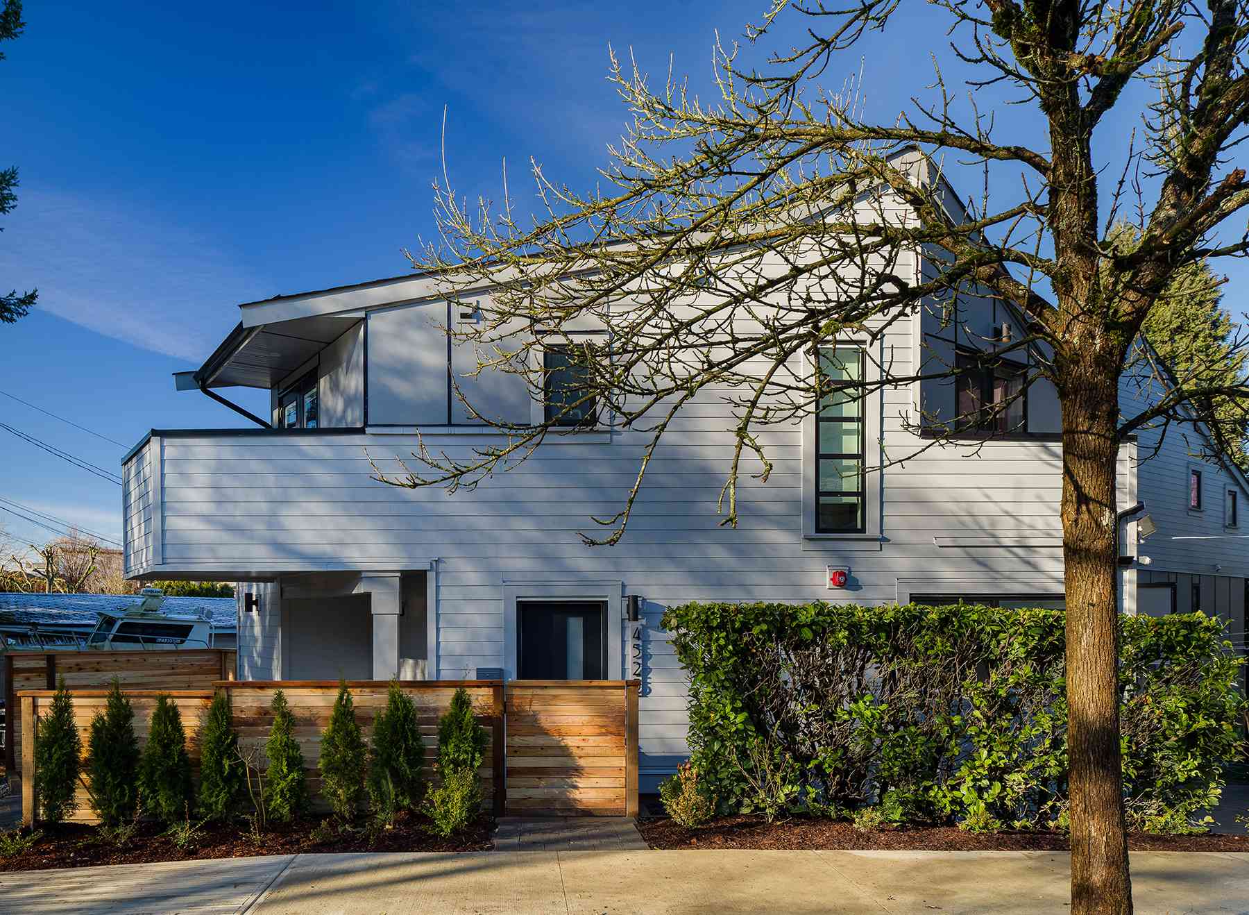 The Park Residences – A contemporary two-story residential building with white horizontal siding and an upper balcony, viewed from the street in bright daylight. In front, a warm wood slat fence and gate are bordered by young cypress shrubs, a manicured green hedge, and a bare tree in the foreground under a clear blue sky.