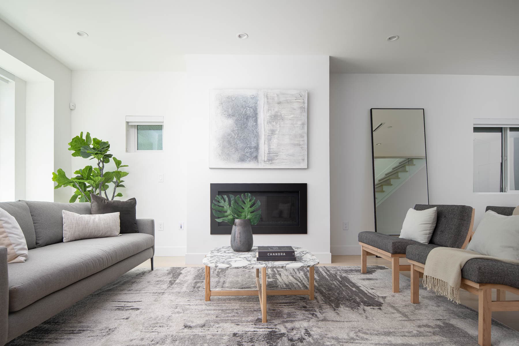 The Park Residences – A bright and modern living room featuring a recessed black fireplace beneath an abstract canvas painting on a clean white wall. The seating area includes a gray upholstered sofa, two wooden-framed accent chairs, a marble coffee table, and an abstract-patterned area rug, accented by indoor potted greenery and a tall leaning mirror.