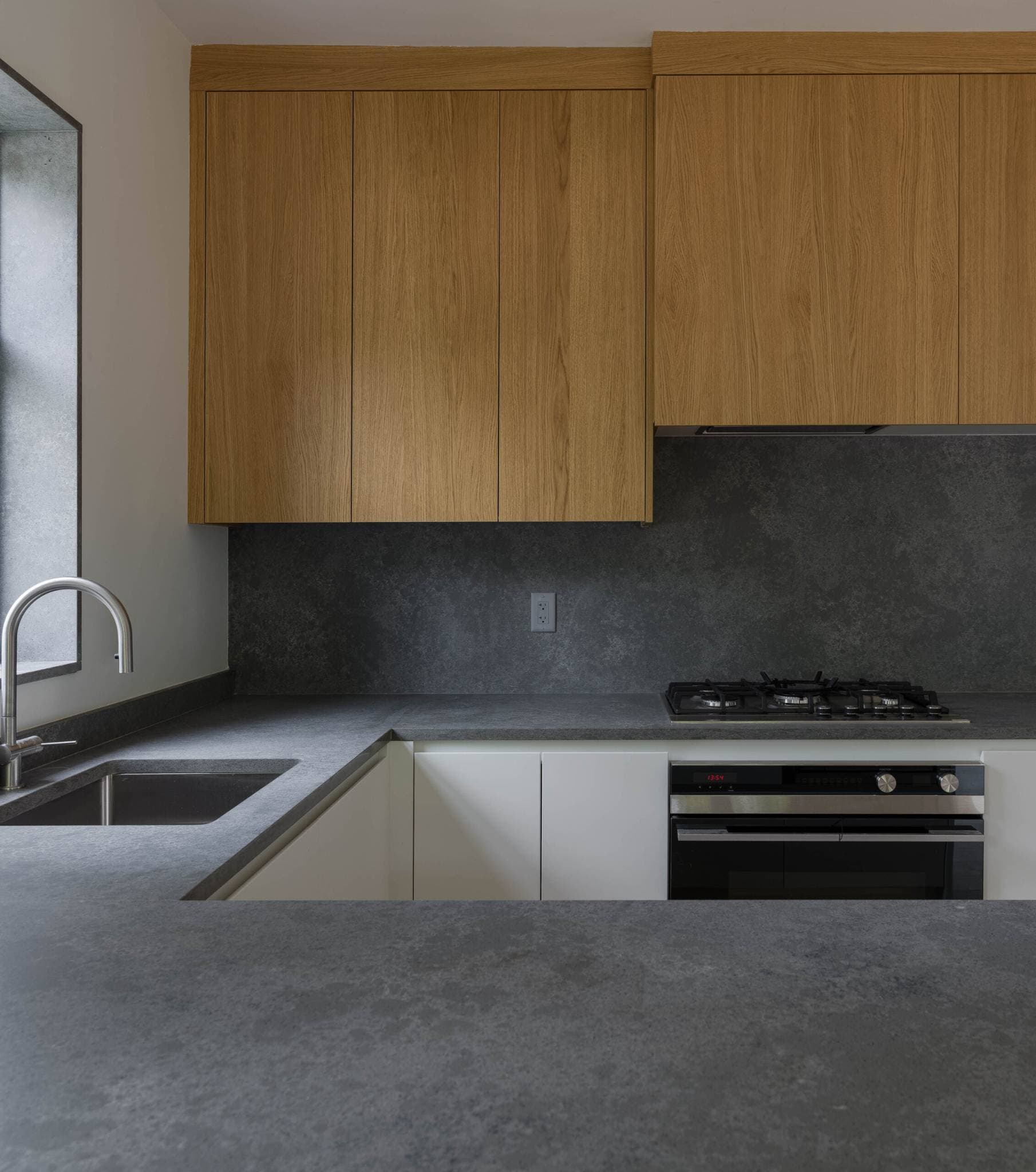 The Park Residences – A modern kitchen detail featuring matte dark gray stone countertops and matching full-height backsplash paired with light wood upper cabinets and white lower cabinetry. An undermount stainless steel sink with a curved chrome faucet sits beside a window opening, alongside an integrated gas cooktop and built-in oven.