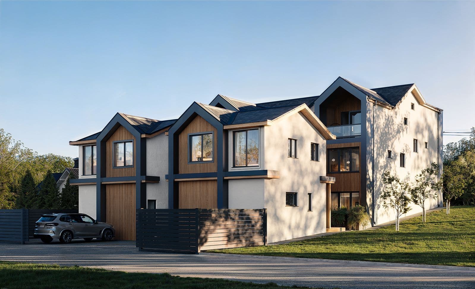 Solin – A modern multi-unit residential development with gabled rooflines, featuring white stucco walls, dark blue trim, and warm wood accent siding. A silver SUV is parked on the paved driveway in front of dark horizontal slatted fences, while green grass and trees surround the building under a clear blue sky.