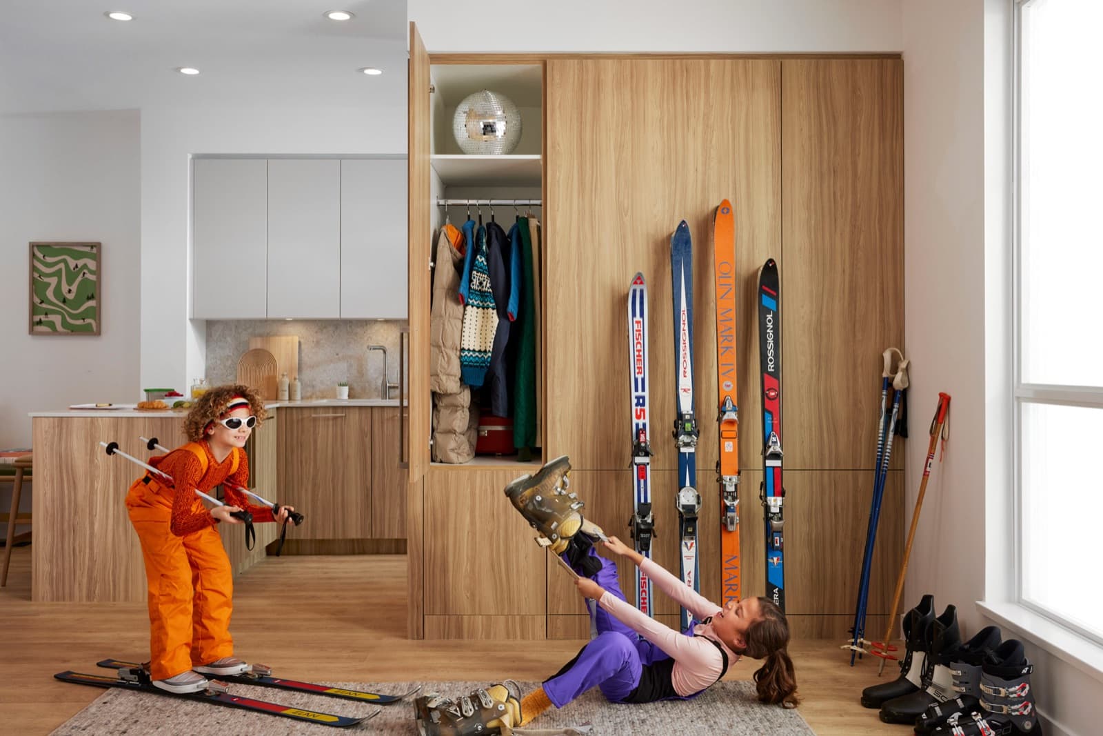 Towns at Lynn – Mudroom-style storage closet with skis, boots, and coats, next to two children dressed in ski gear near an open kitchen.
