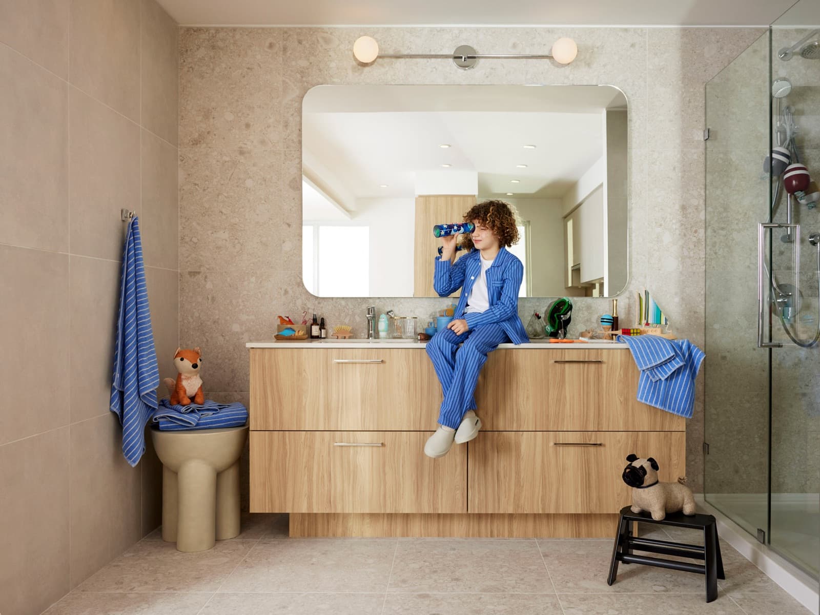 Towns at Lynn – Bathroom with a wood double vanity, glass shower, and a boy sitting on the counter drinking from a water bottle.