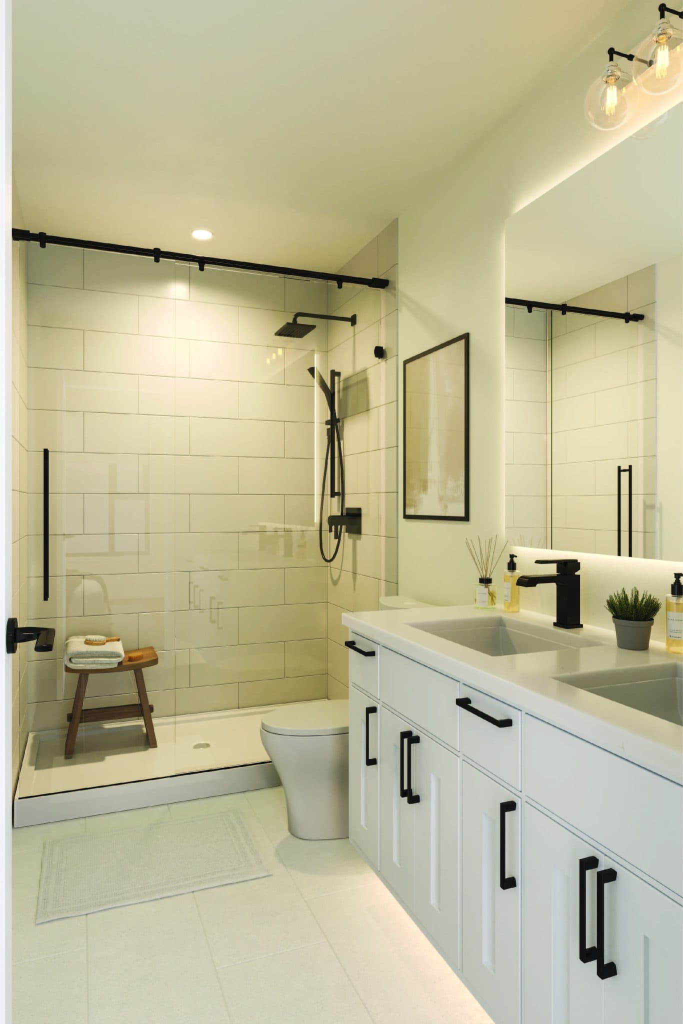 Chronicle – A modern, bright bathroom featuring a white double-sink vanity with matte black hardware, an illuminated mirror, and a toilet. A glass-enclosed walk-in shower is lined with off-white subway tile and equipped with matte black fixtures and a small wooden stool.