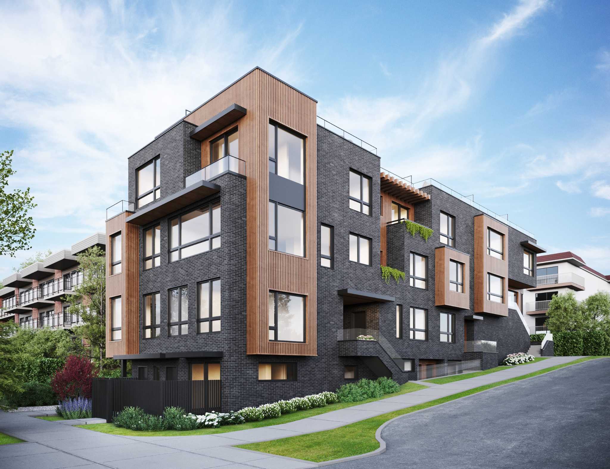 The Vantage – A modern multi-story residential building clad in dark charcoal brick and vertical natural wood accents sits along a sloping suburban street. Large windows, glass-railed balconies, and landscaped curbside greenery define the facade under a clear daylight sky.