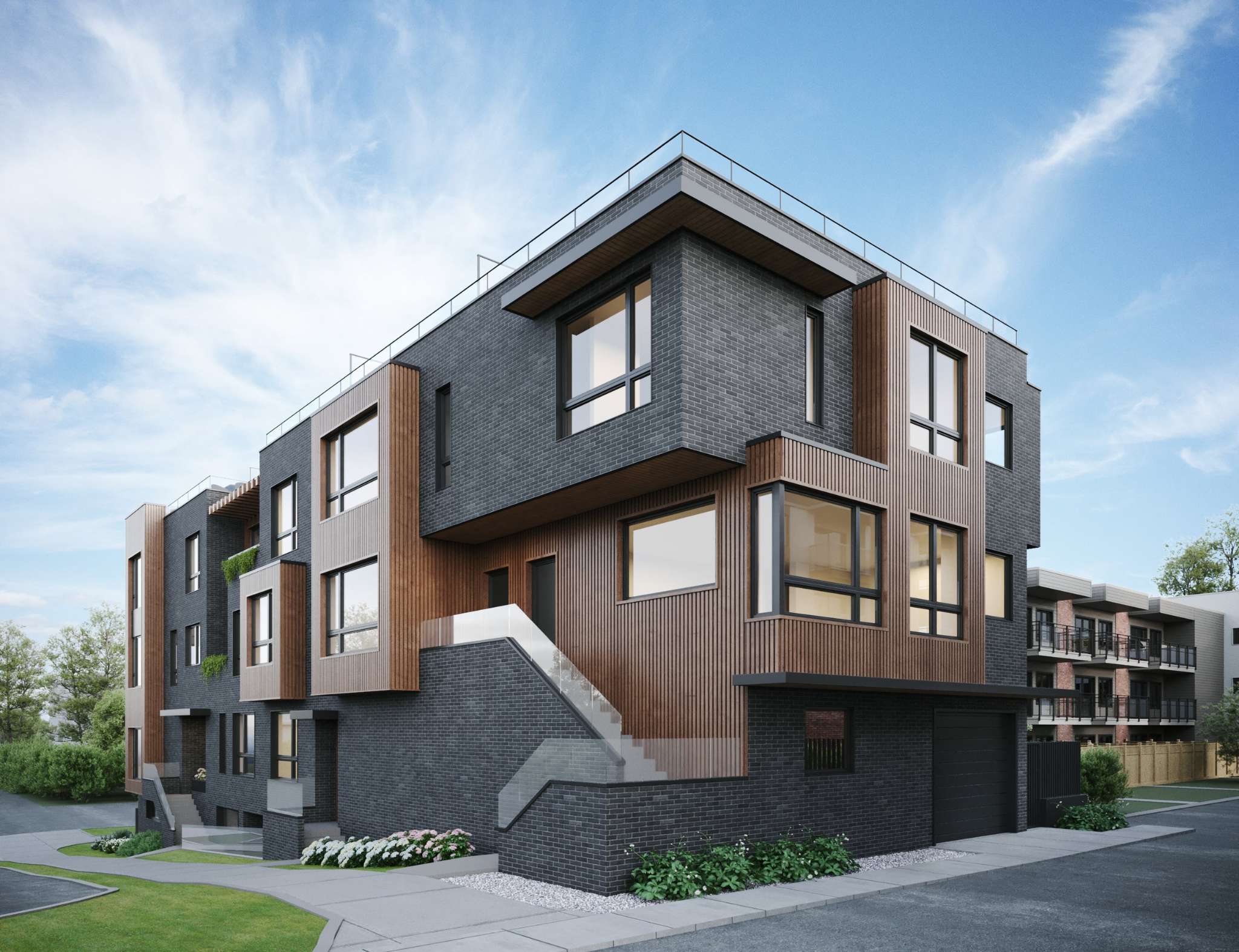 The Vantage – A contemporary multi-level residential building features charcoal grey brick paired with vertical warm wood slat paneling and black-framed windows. An exterior staircase with glass railings leads up to the main entrance above a ground-level garage, all framed by concrete walkways, manicured greenery, and a bright blue sky.