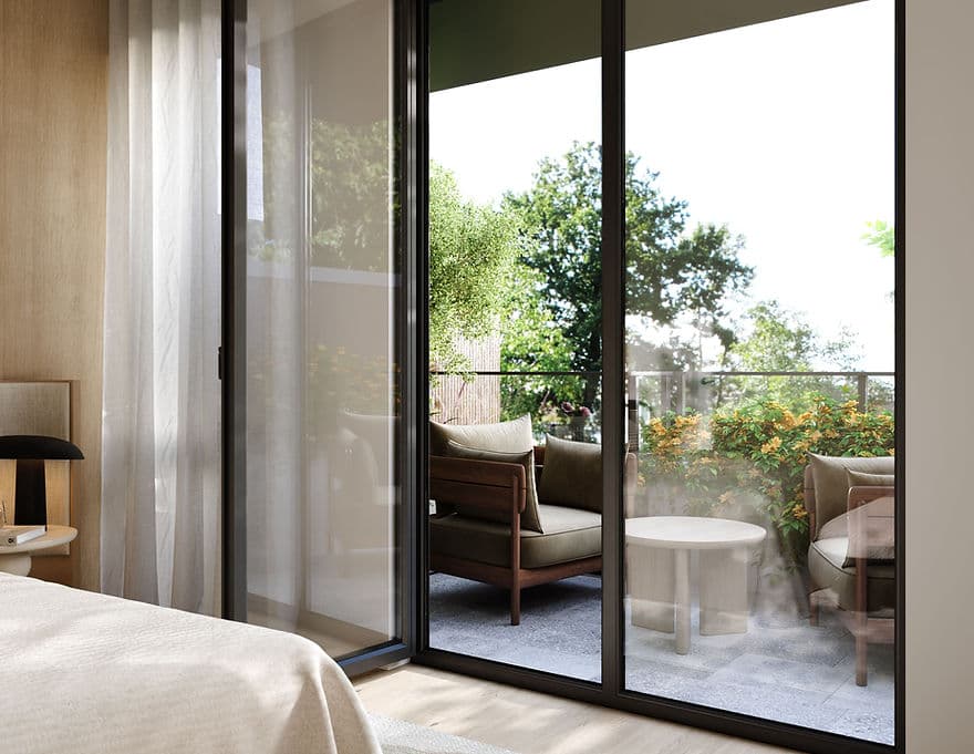 Park & Granville – A view looking out from a bedroom through large glass sliding doors onto a private patio or balcony furnished with wooden armchairs, a round coffee table, and lush flowering shrubs. Sunlight filters into the serene room, highlighting sheer curtains, a soft bedspread, and leafy green trees visible just beyond the glass railing.
