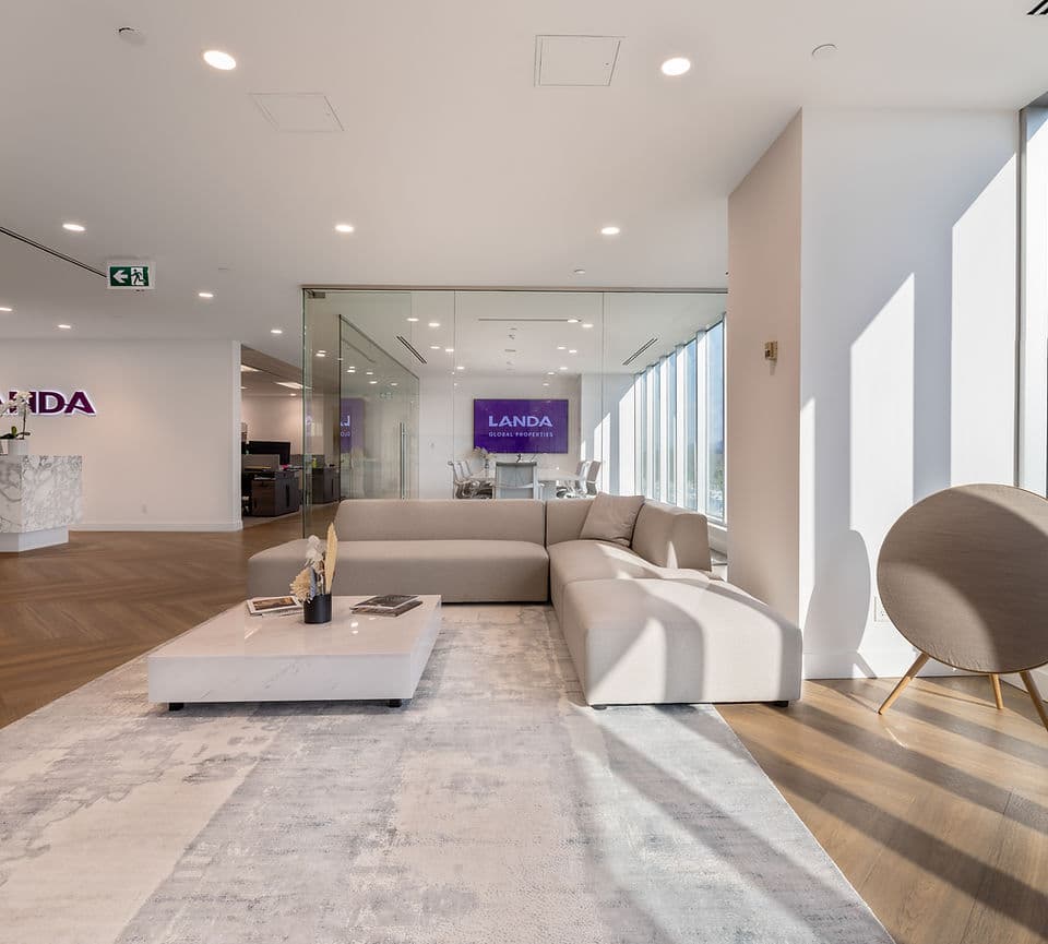 Park & Granville – A bright, modern commercial office reception and lounge area featuring a large beige sectional sofa, a white marble coffee table, and an area rug over herringbone wood floors. Floor-to-ceiling windows cast natural daylight into the space, with a glass-enclosed conference room visible in the background.