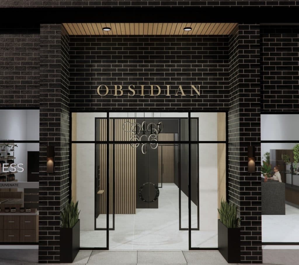 Obsidian on Tenth – A ground-level view of a modern building entrance framed by dark charcoal brickwork and warm wood paneling under the soffit, with gold-toned lettering reading 'OBSIDIAN' above the door. Large glass doors and flanking windows reveal a brightly lit lobby featuring light stone flooring, vertical wood slat accent walls, and a contemporary circular ring chandelier.