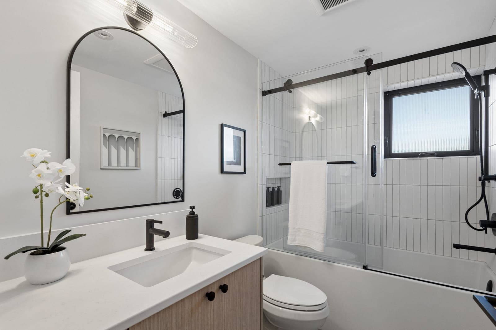 Victoria Haus – A modern bathroom featuring a light wood vanity with a white countertop, an arched black-framed mirror, and matte black fixtures. Adjacent is a combined bathtub and shower enclosed by sliding glass doors, lined with vertical white subway tiles and equipped with black hardware.