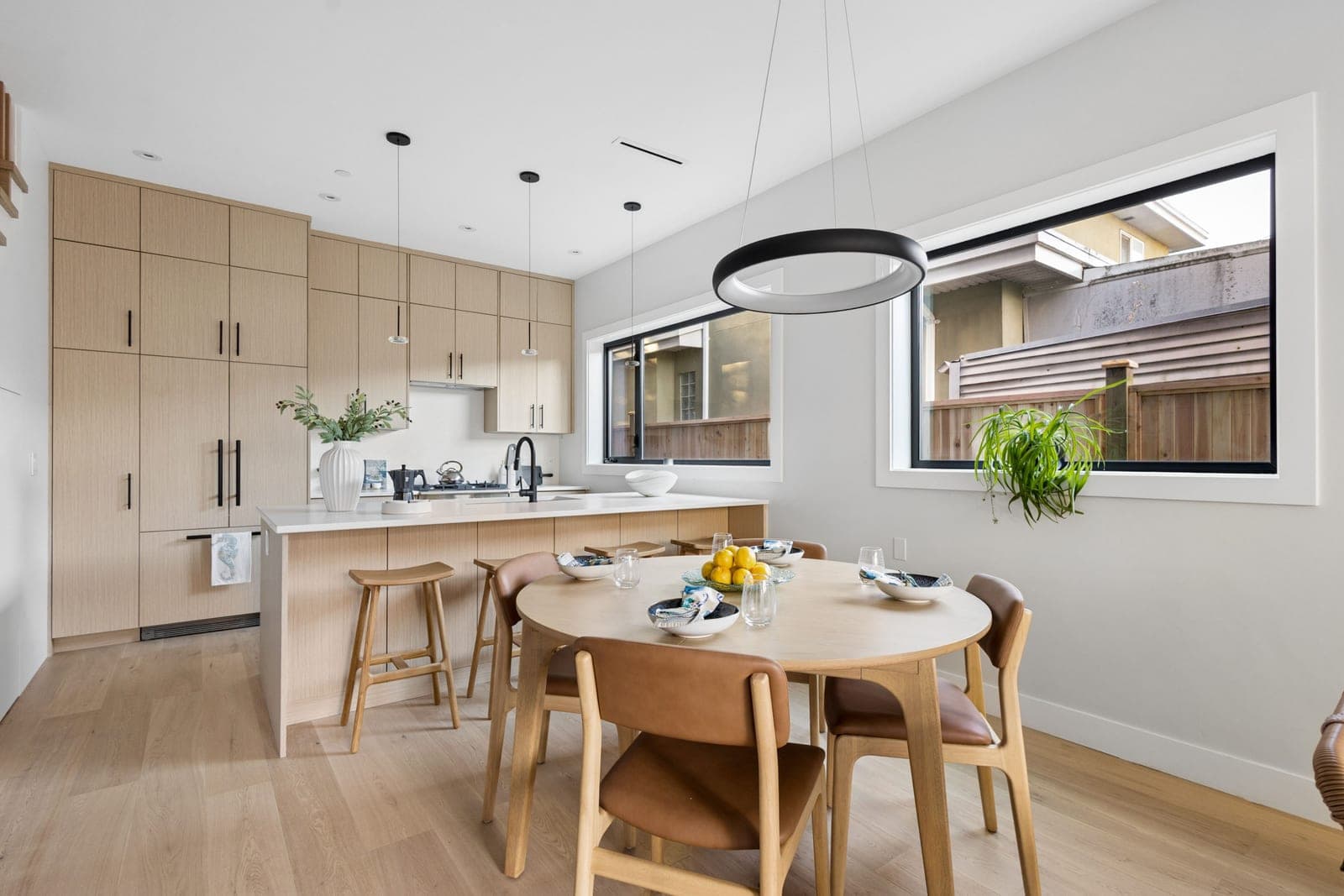 Victoria Haus – A bright, contemporary open-concept kitchen and dining area featuring light wood flat-panel cabinetry, white countertops, and light hardwood floors. In the foreground, a round wooden dining table with leather-seated chairs sits beneath a modern halo chandelier, while wide windows let in ample natural light.